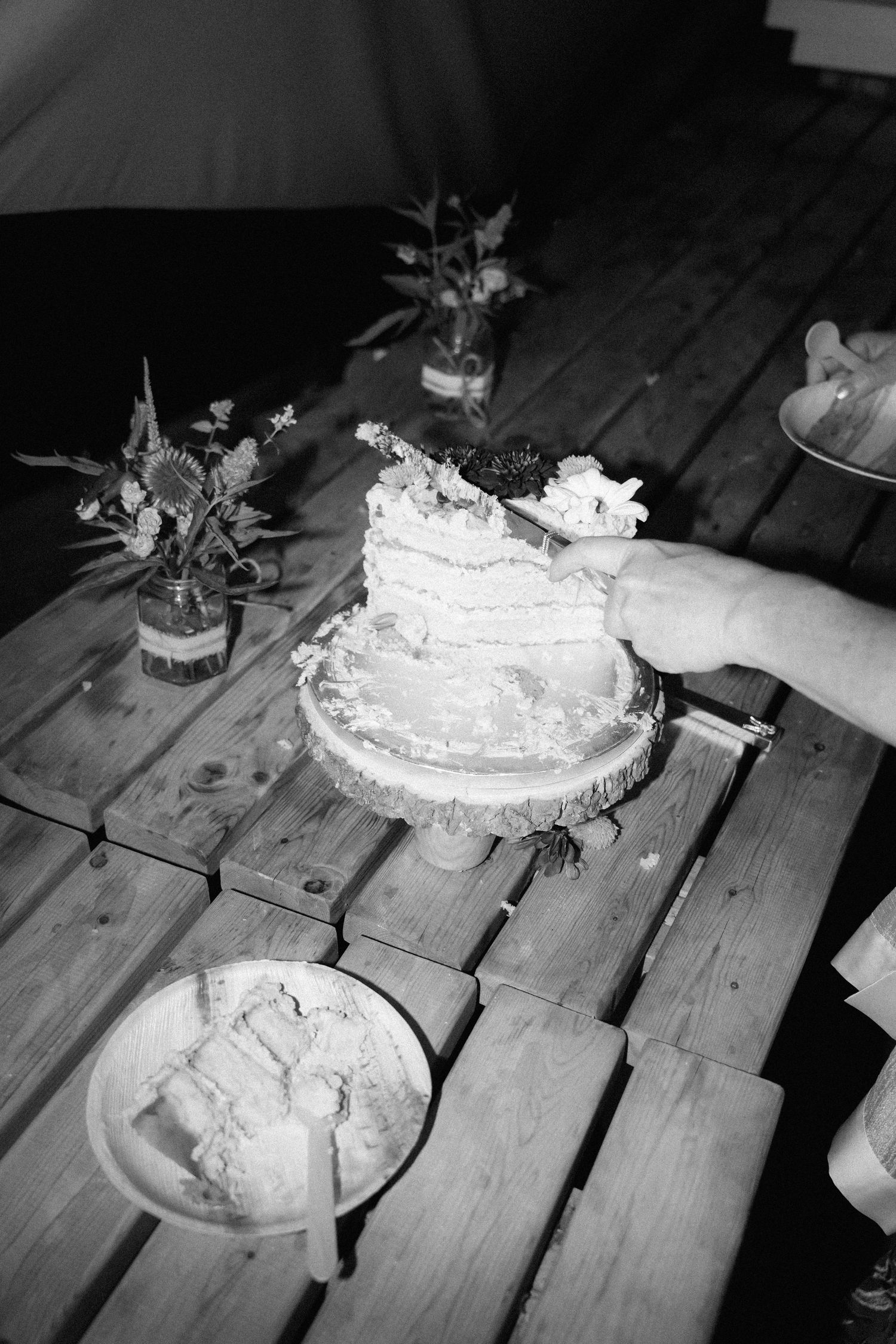 Black and white photograph of a messy layered cake being cut on a wooden table with small vases of flowers nearby.