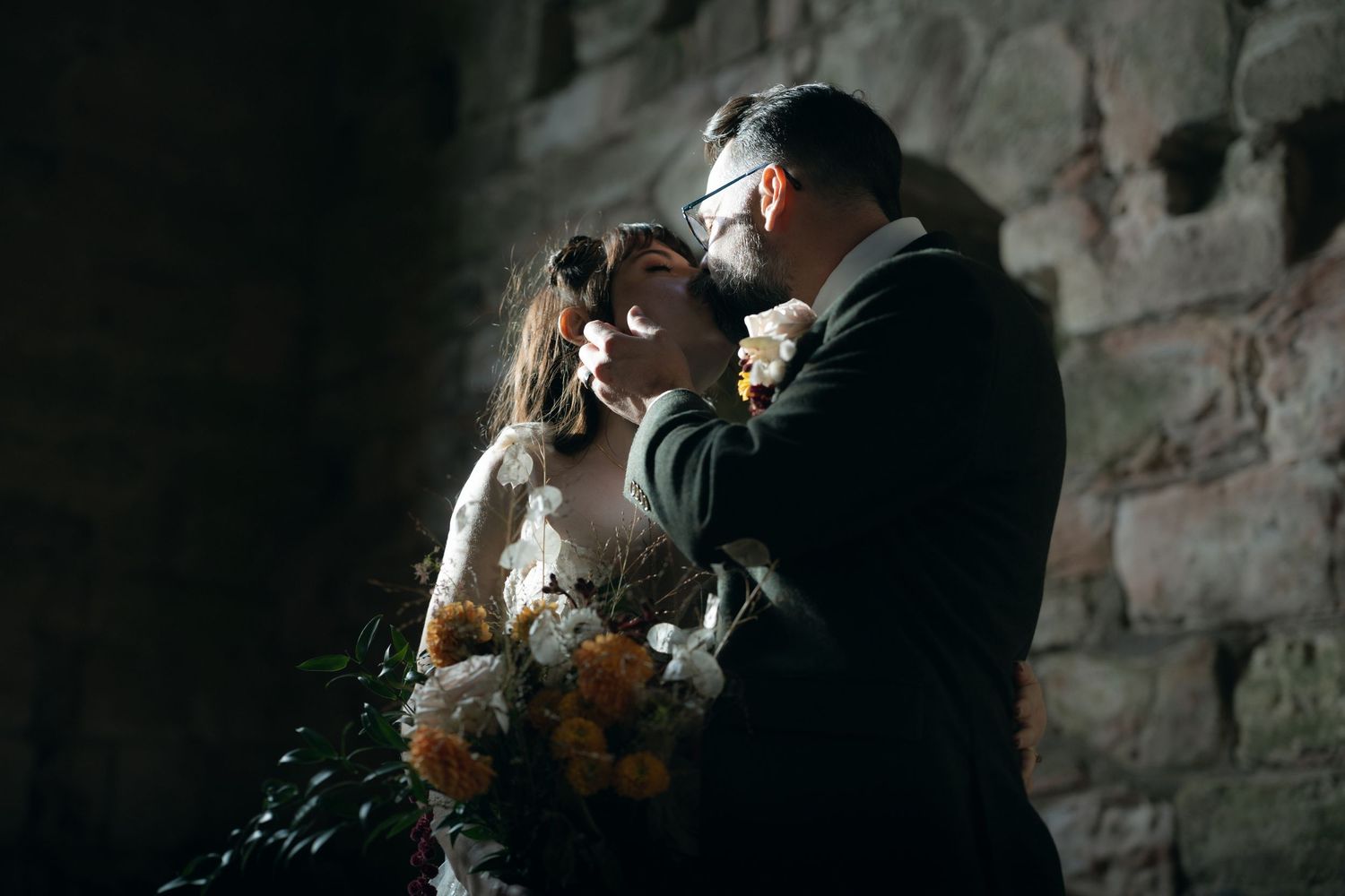 A romantic wedding couple embraces and kisses in a rustic stone setting with dramatic lighting and shadows.