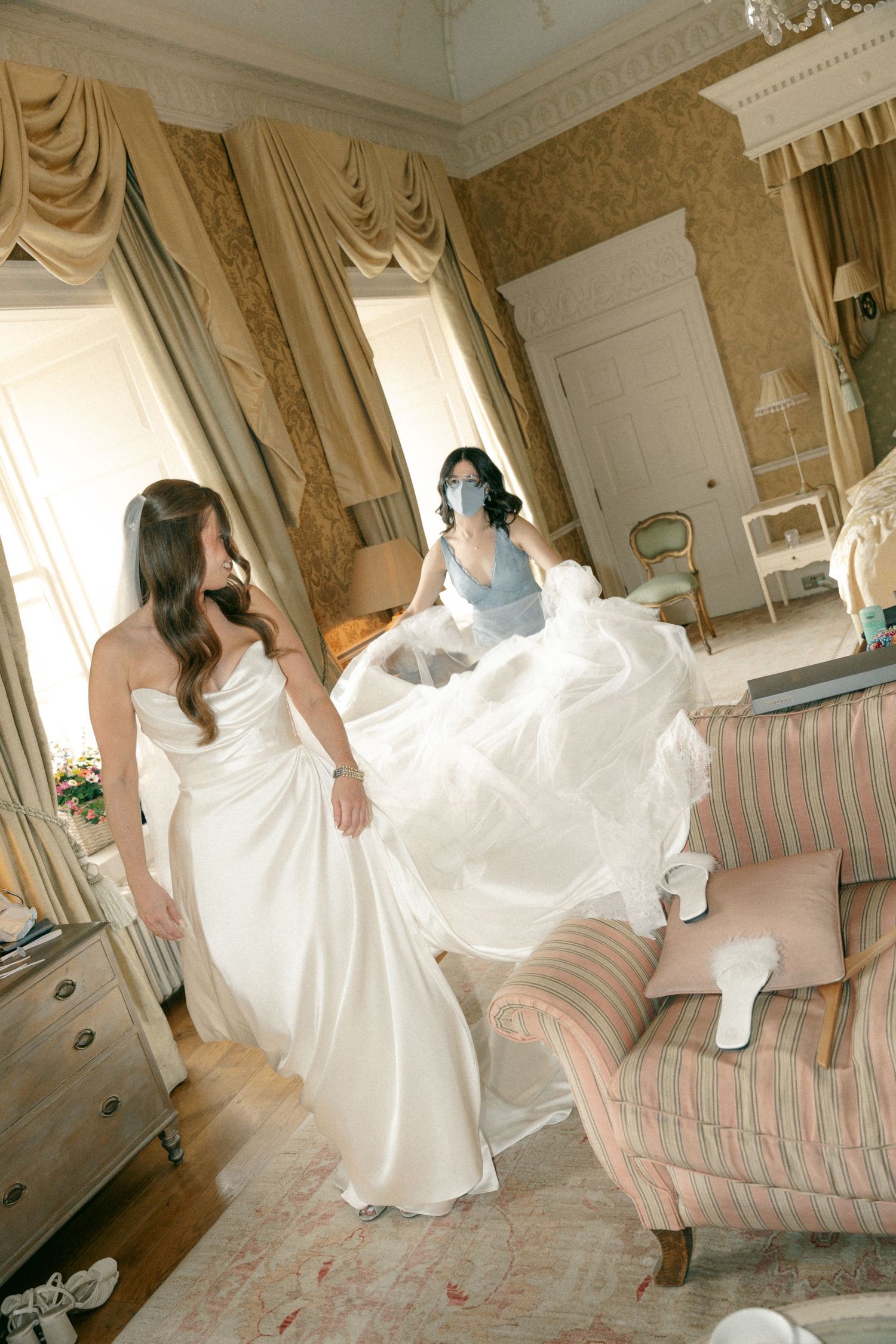 A bride in a flowing white wedding gown twirls playfully in an elegant room with pink striped furniture and ornate decor.