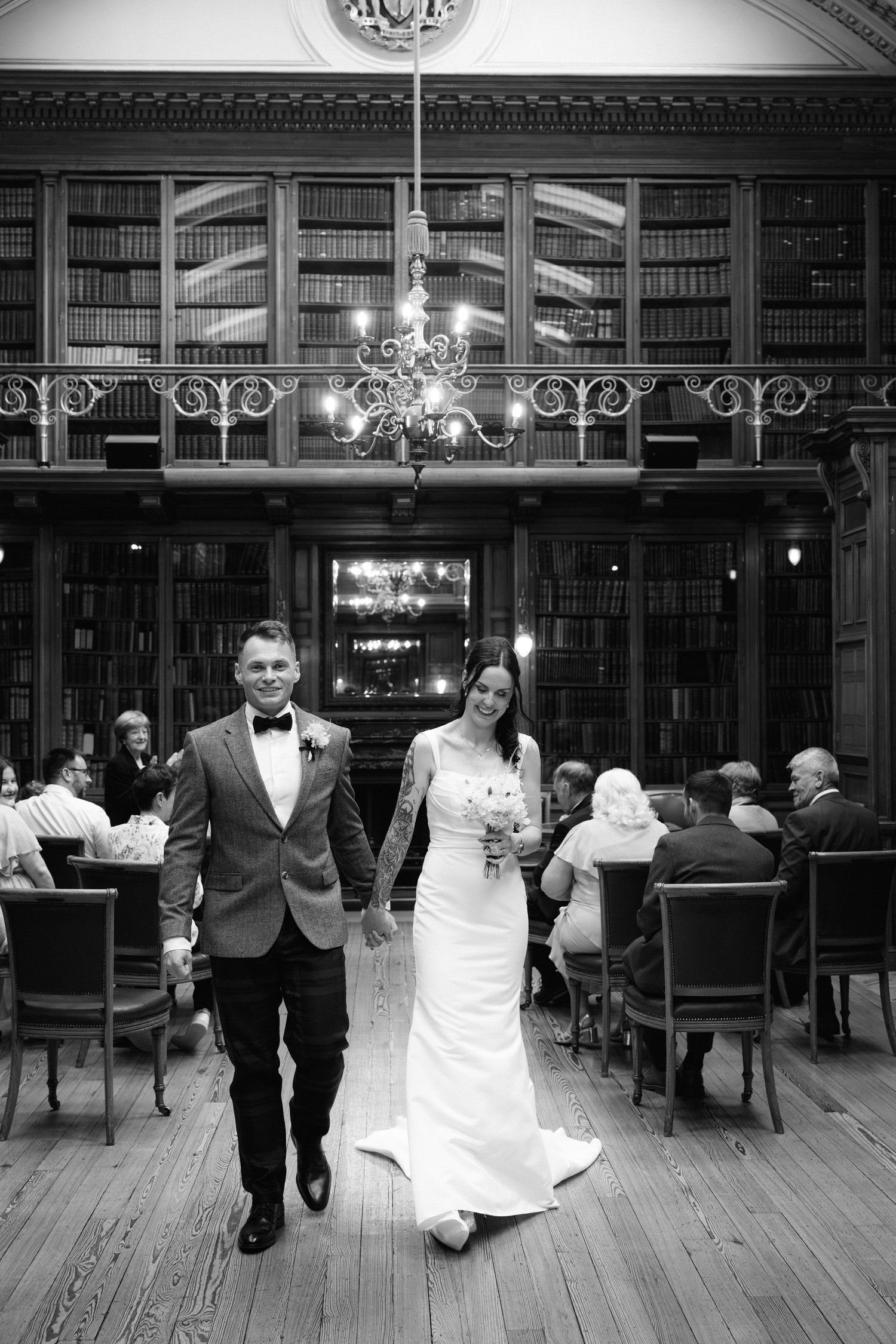 A couple walks down the aisle in a grand library with ornate bookshelves and a chandelier overhead.