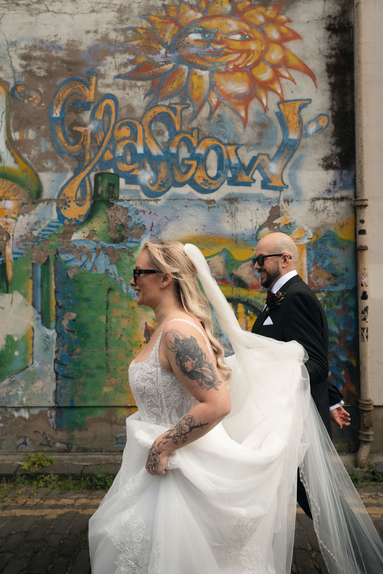 Bride and groom walk past colorful Glasgow graffiti mural, bride in lace gown with veil, groom in black suit.