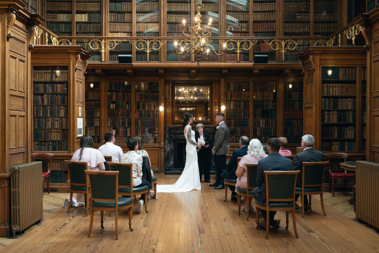 Wedding ceremony taking place in an elegant vintage library with wooden bookshelves and chandeliers.