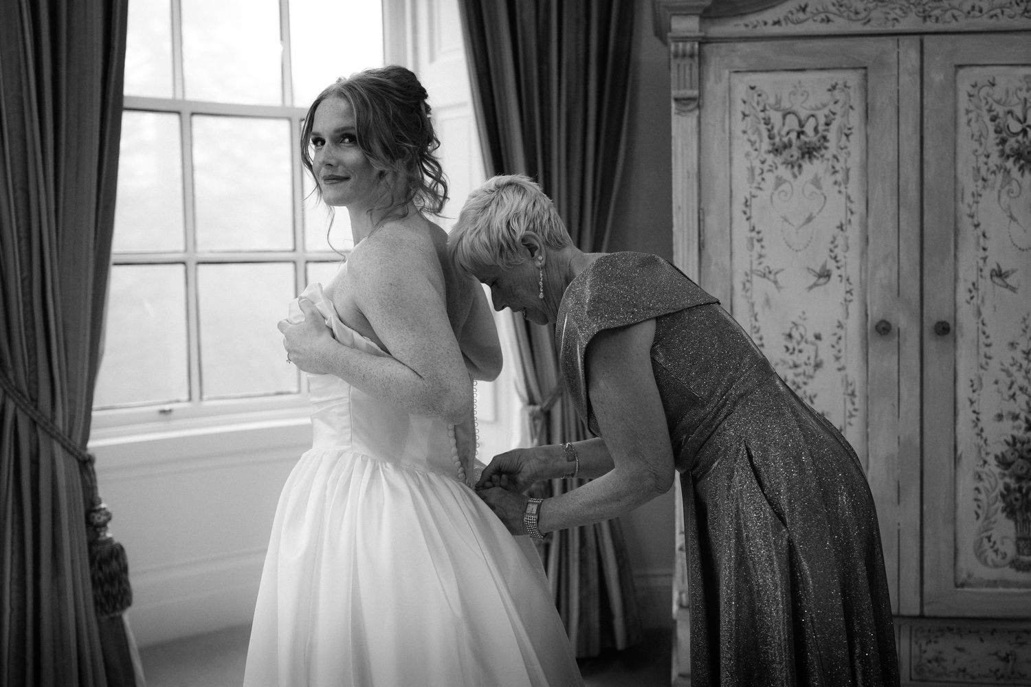 A bride in a white wedding dress gets ready in an elegant room with vintage decor and natural window light.