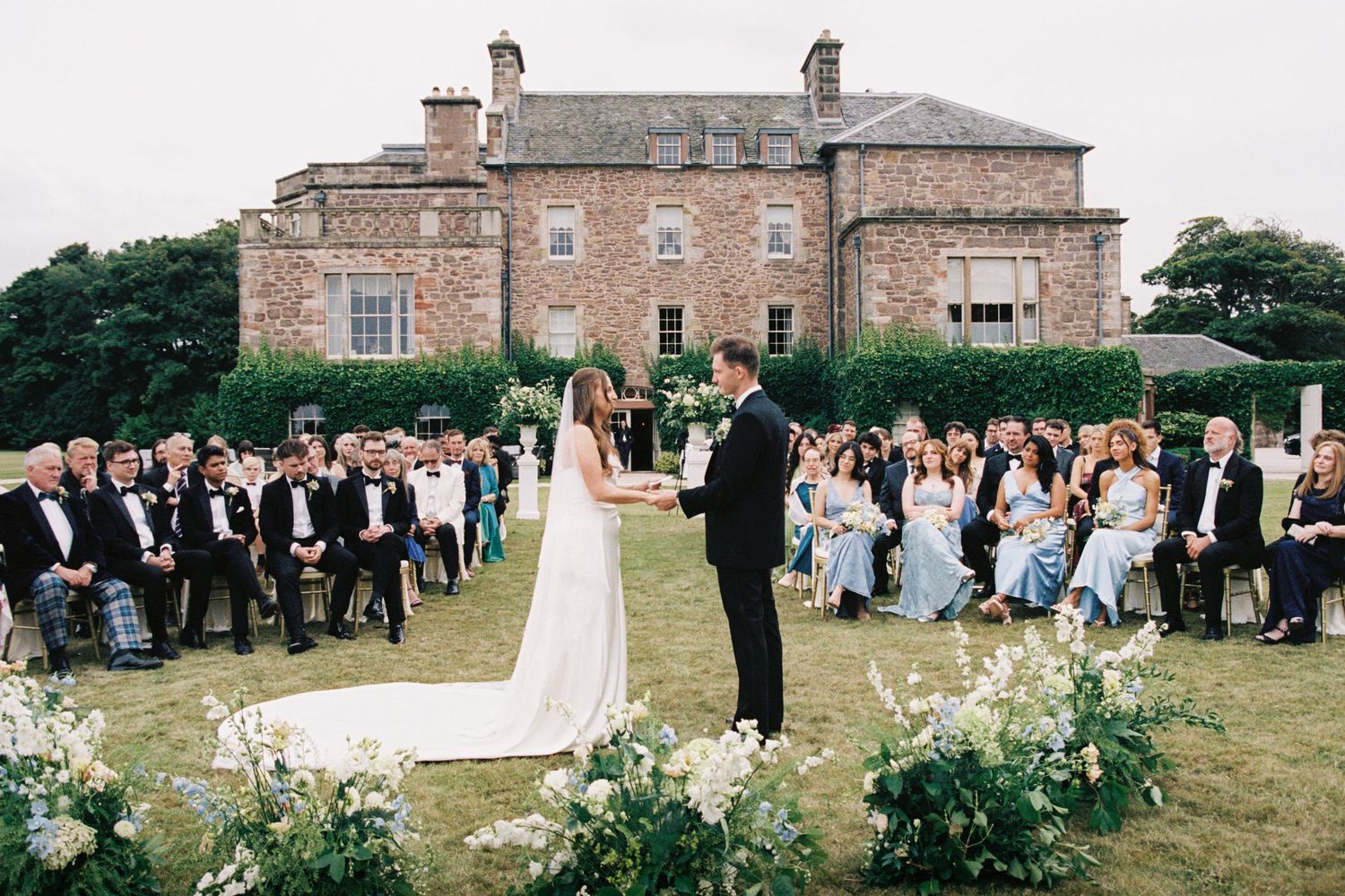 Panoramic view of an elegant outdoor wedding ceremony at a historic stone mansion with guests seated on the lawn.