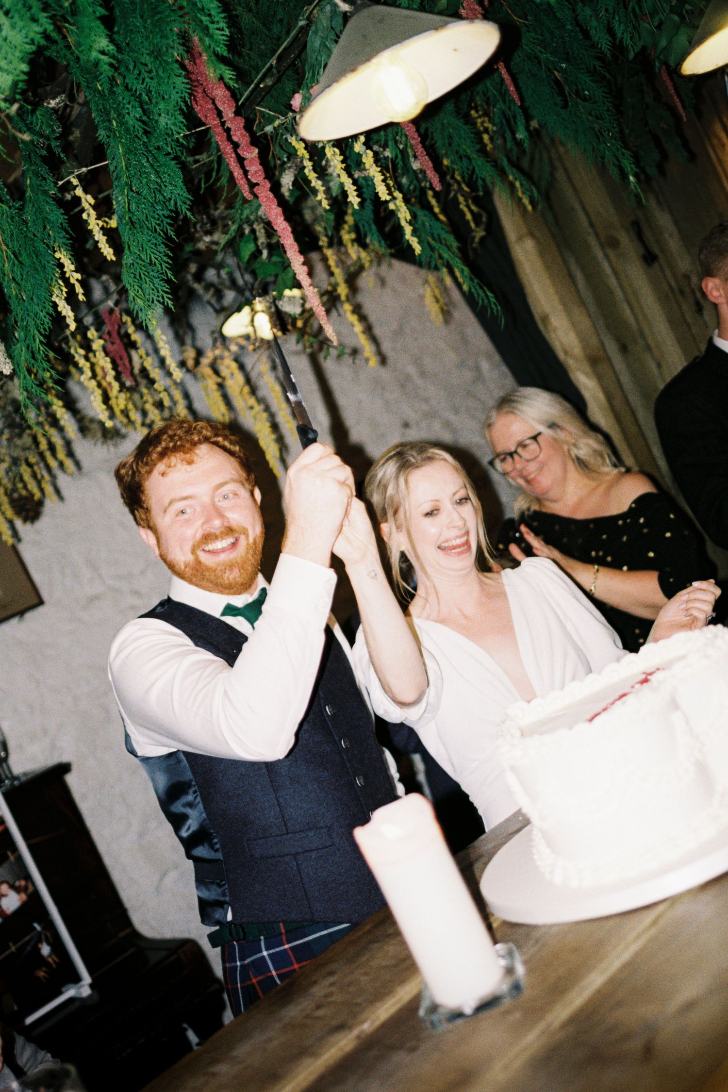 A couple dances joyfully together at a wedding reception under decorative greenery and lights.