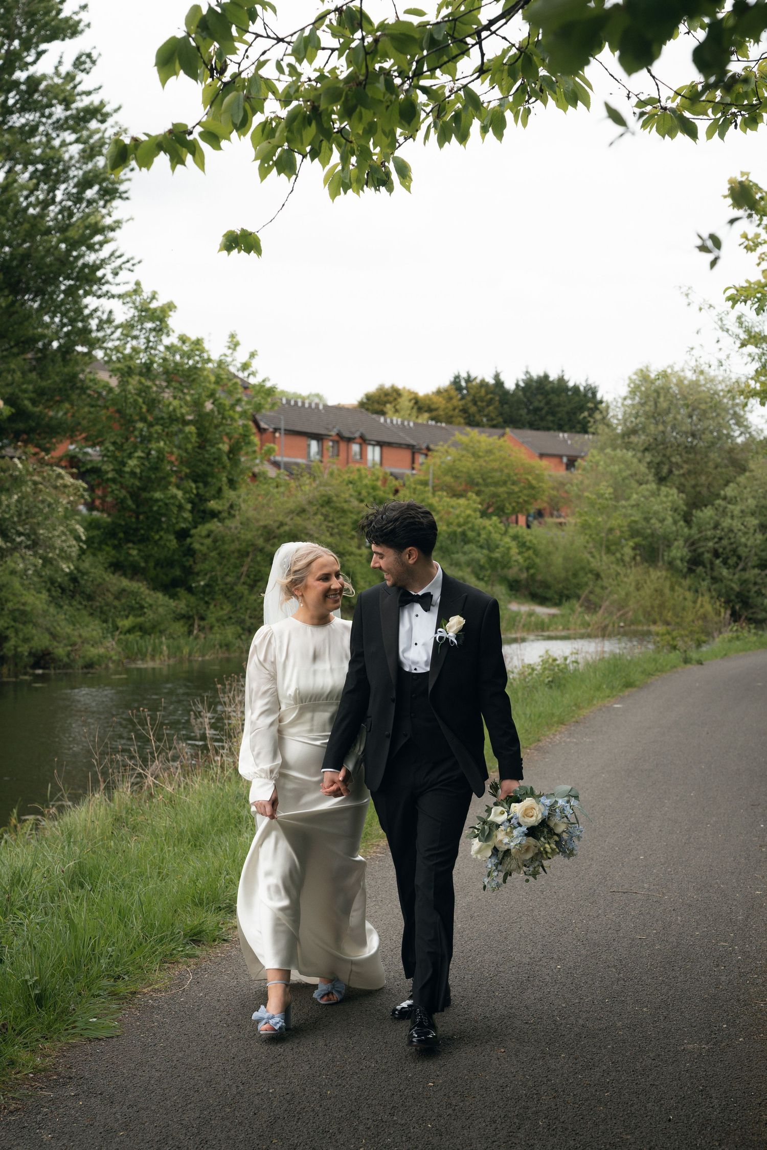 Bride and groom walk hand-in-hand along a canal path, she in white gown, he in black tuxedo holding a bouquet.