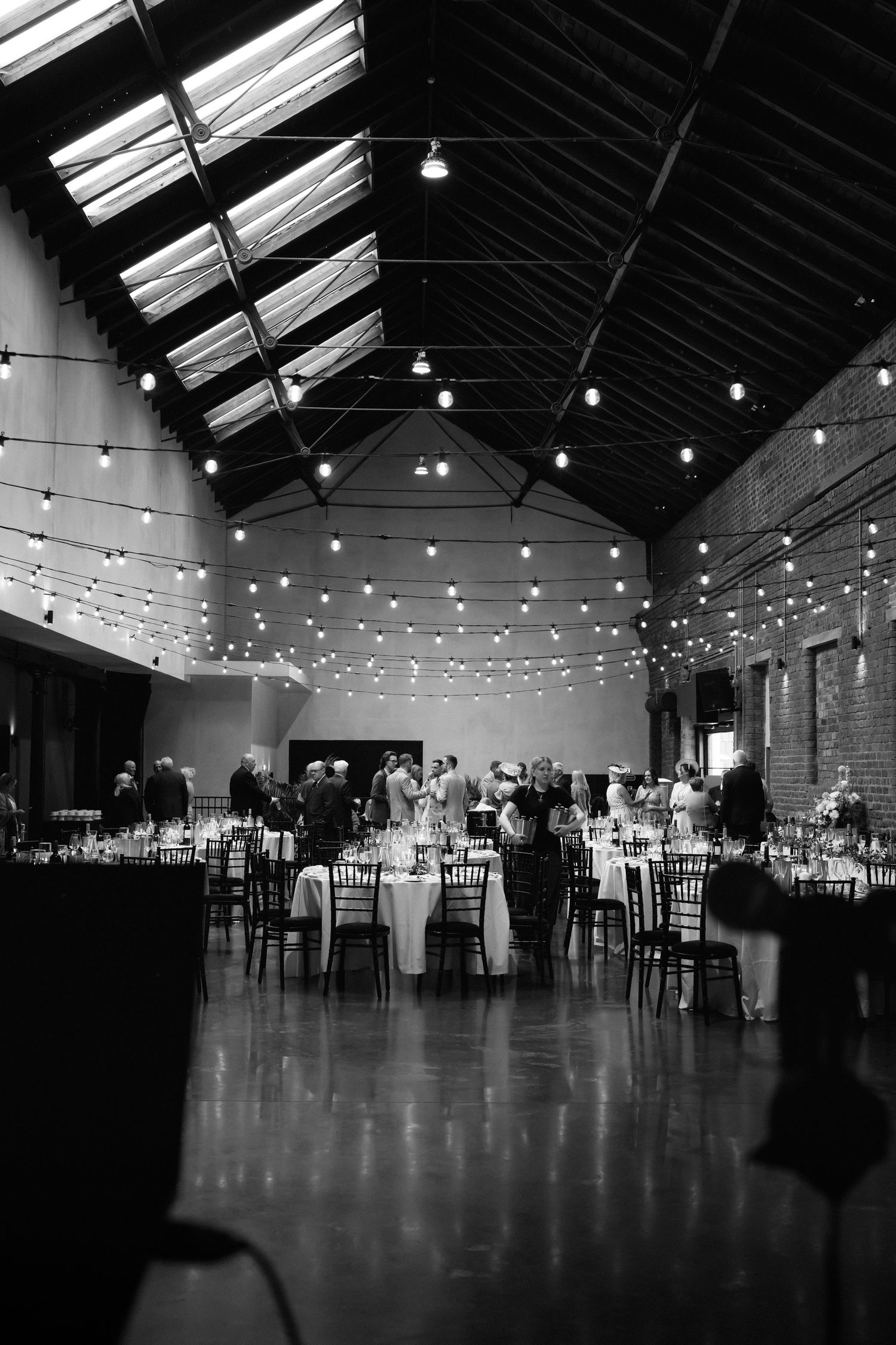Black and white photo of an elegant wedding reception in a rustic venue with string lights and round tables.