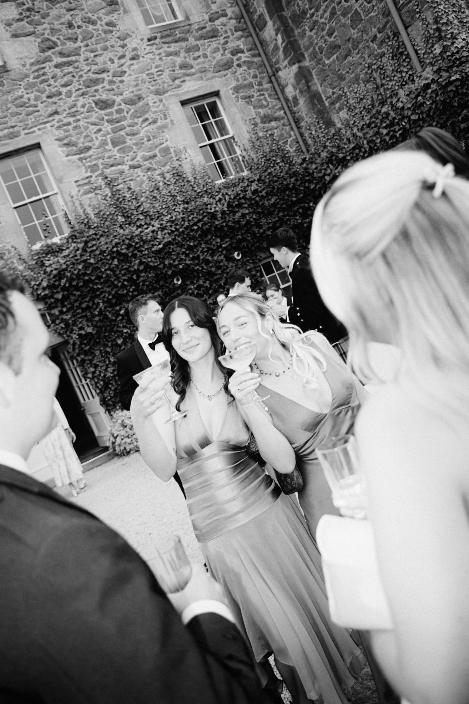 Black and white candid photo of wedding guests laughing together outdoors by a stone building.