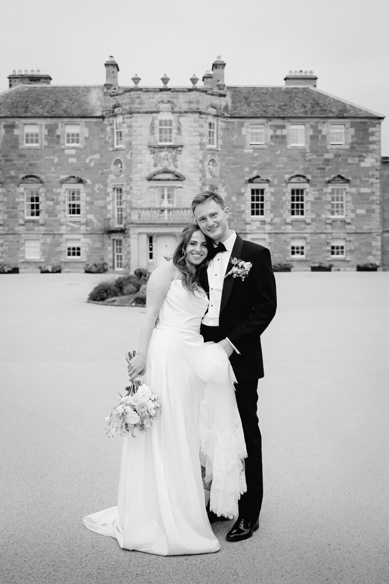 A black and white wedding photo with a couple standing in front of a grand historic stone mansion.