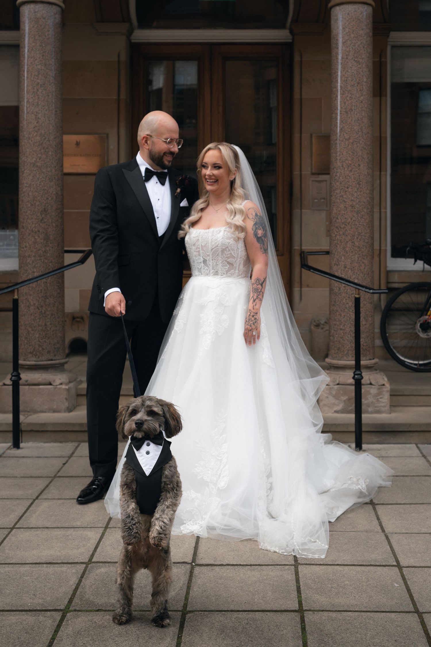 Bride and groom pose with their dog dressed in a tuxedo outside a grand building on their wedding day.