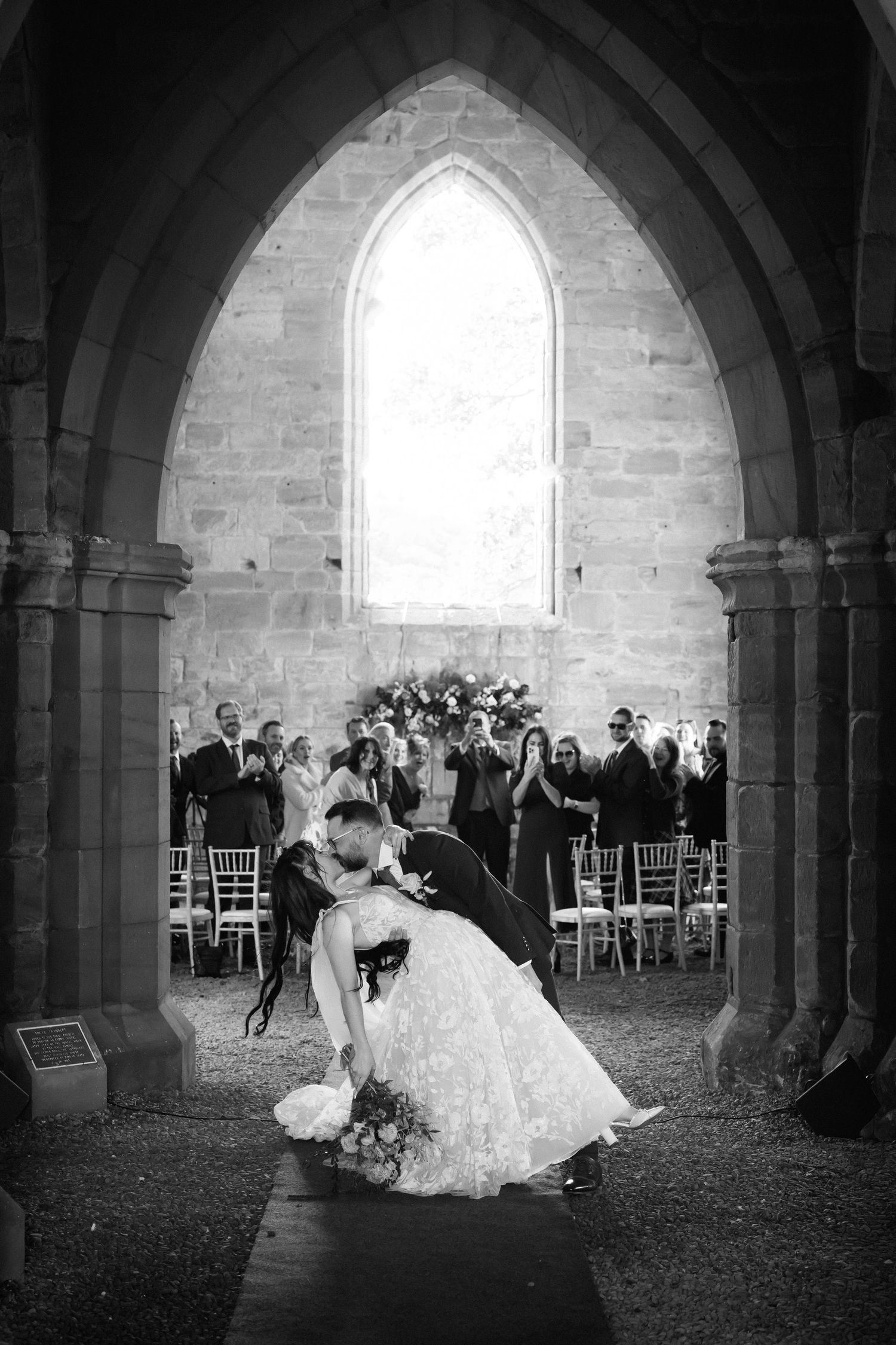 A series of black and white wedding photos showing a couple and guests in a beautiful stone church with arched windows.