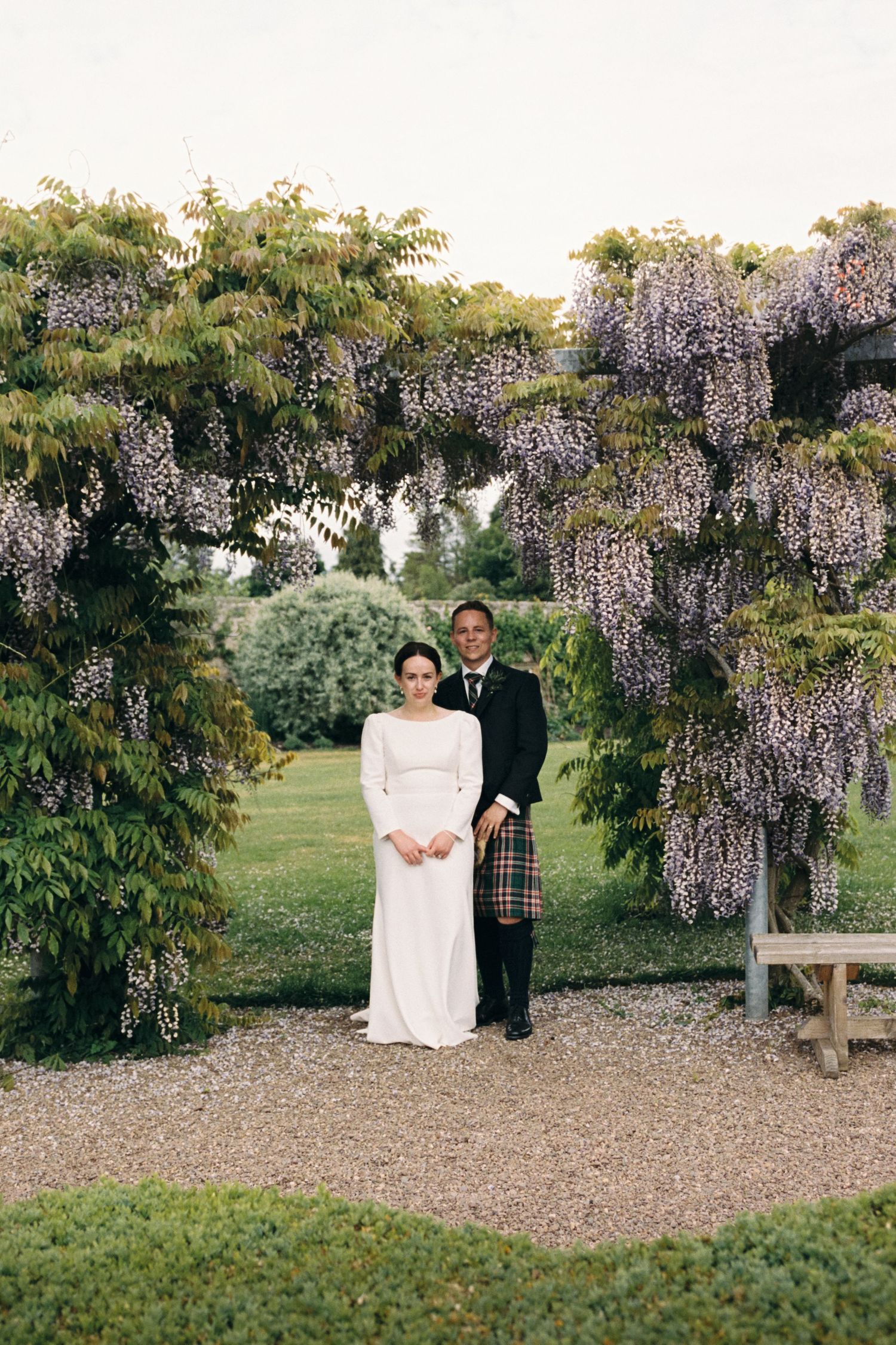 A couple poses beneath blooming wisteria vines in a garden setting during a wedding ceremony.