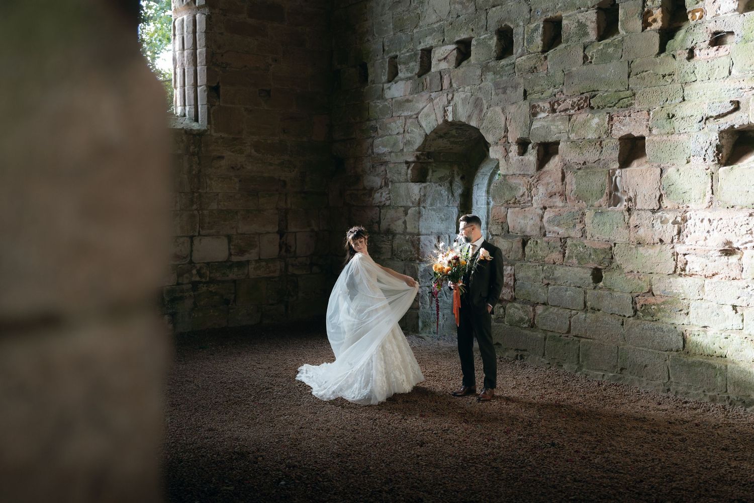 A couple in wedding attire poses together inside a rustic stone building with dramatic lighting.