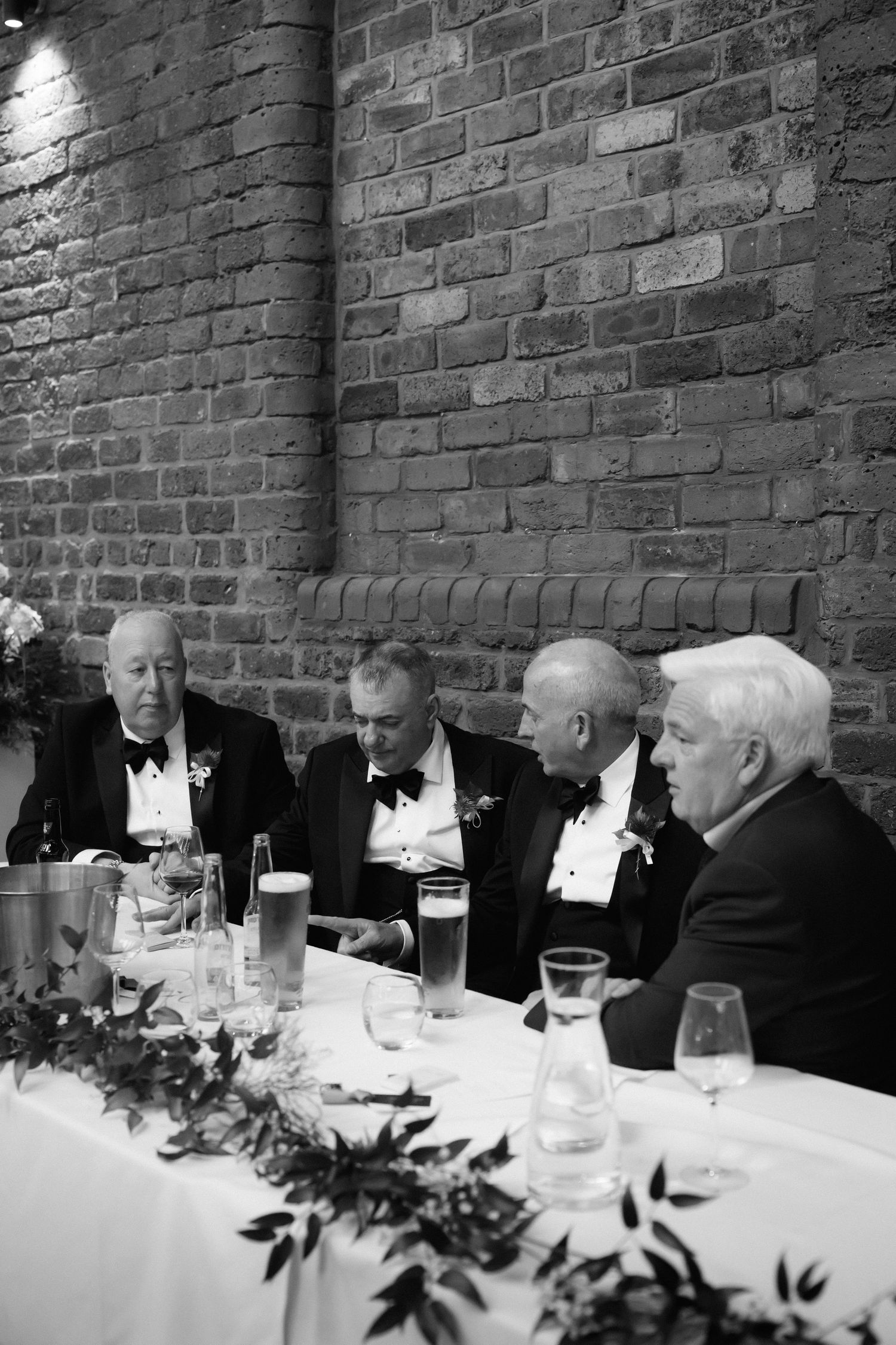 Four men in tuxedos sit at a formal dinner table against a brick wall in this black and white photo.