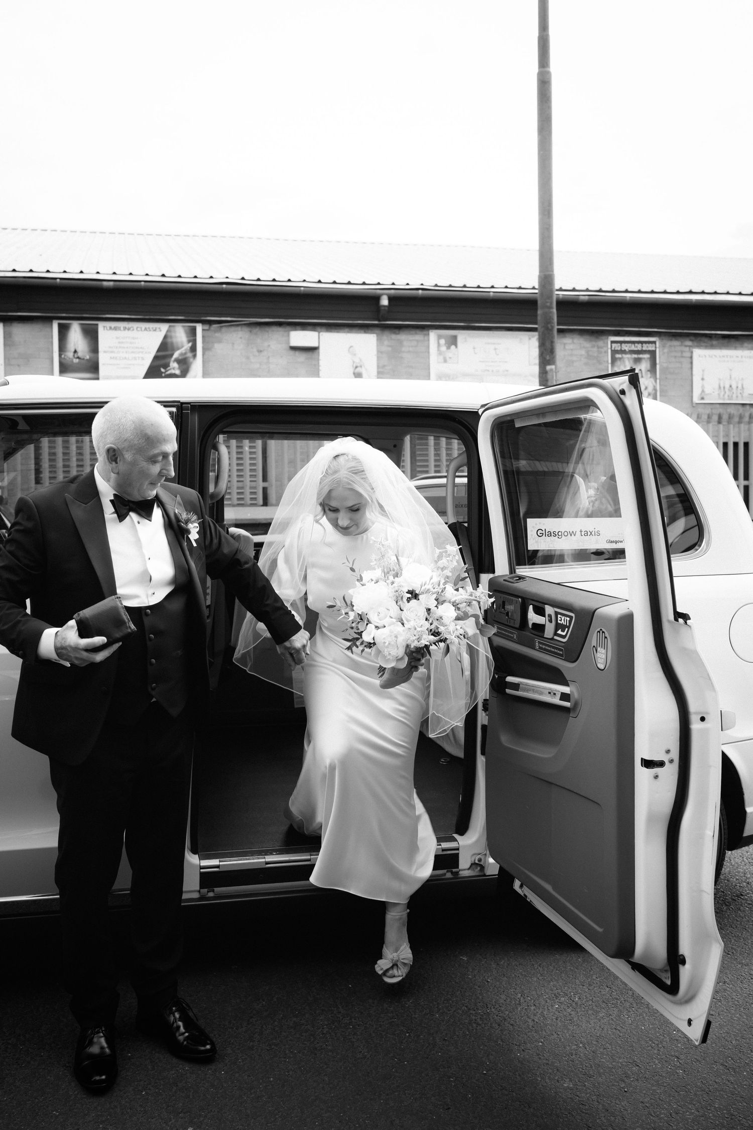 Black and white photo of a bride in a white gown and veil stepping out of a vintage car, assisted by a man in a tuxedo.