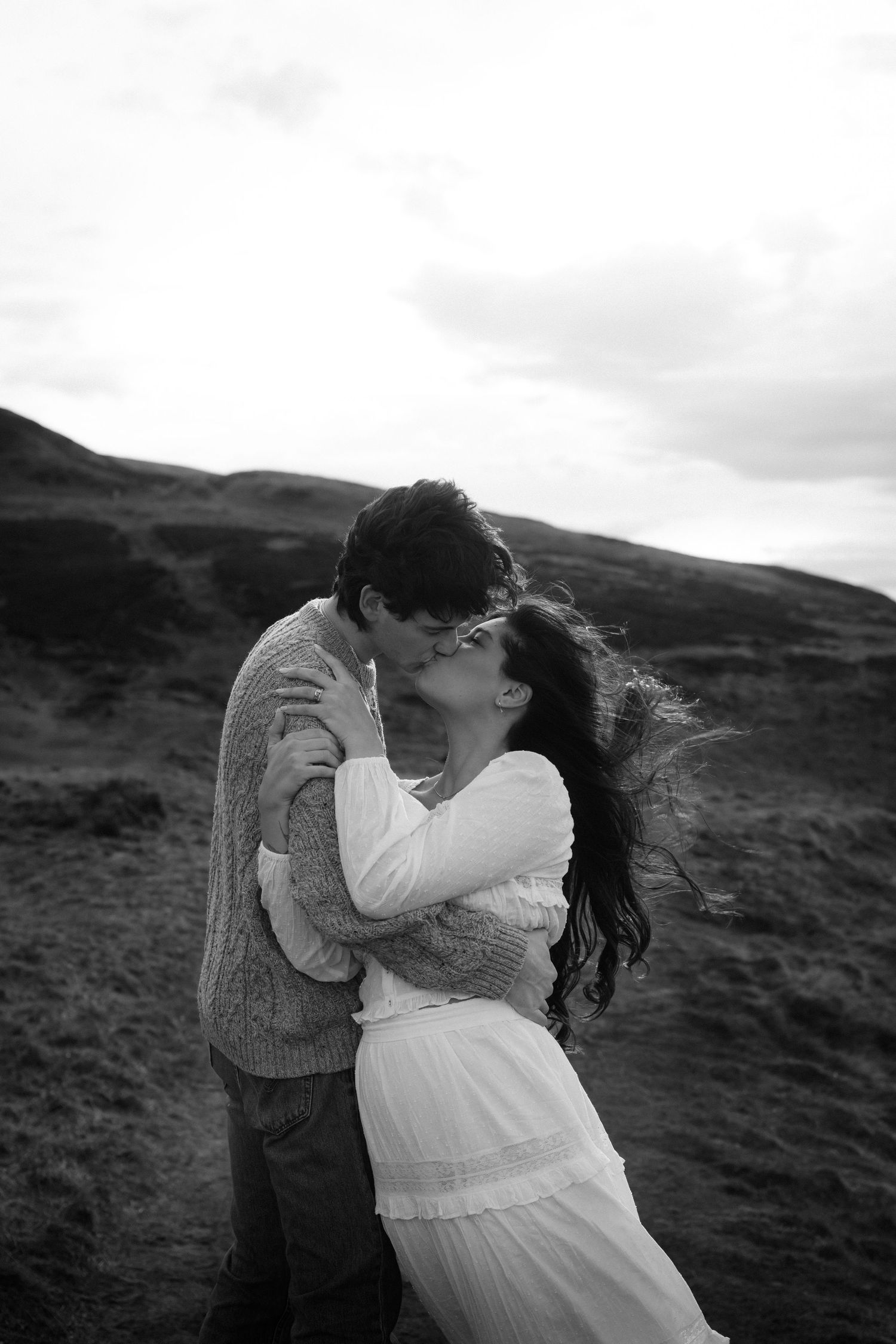 Black and white photo of a couple kissing in a windy highland landscape, woman in white dress, romantic outdoor portrait.