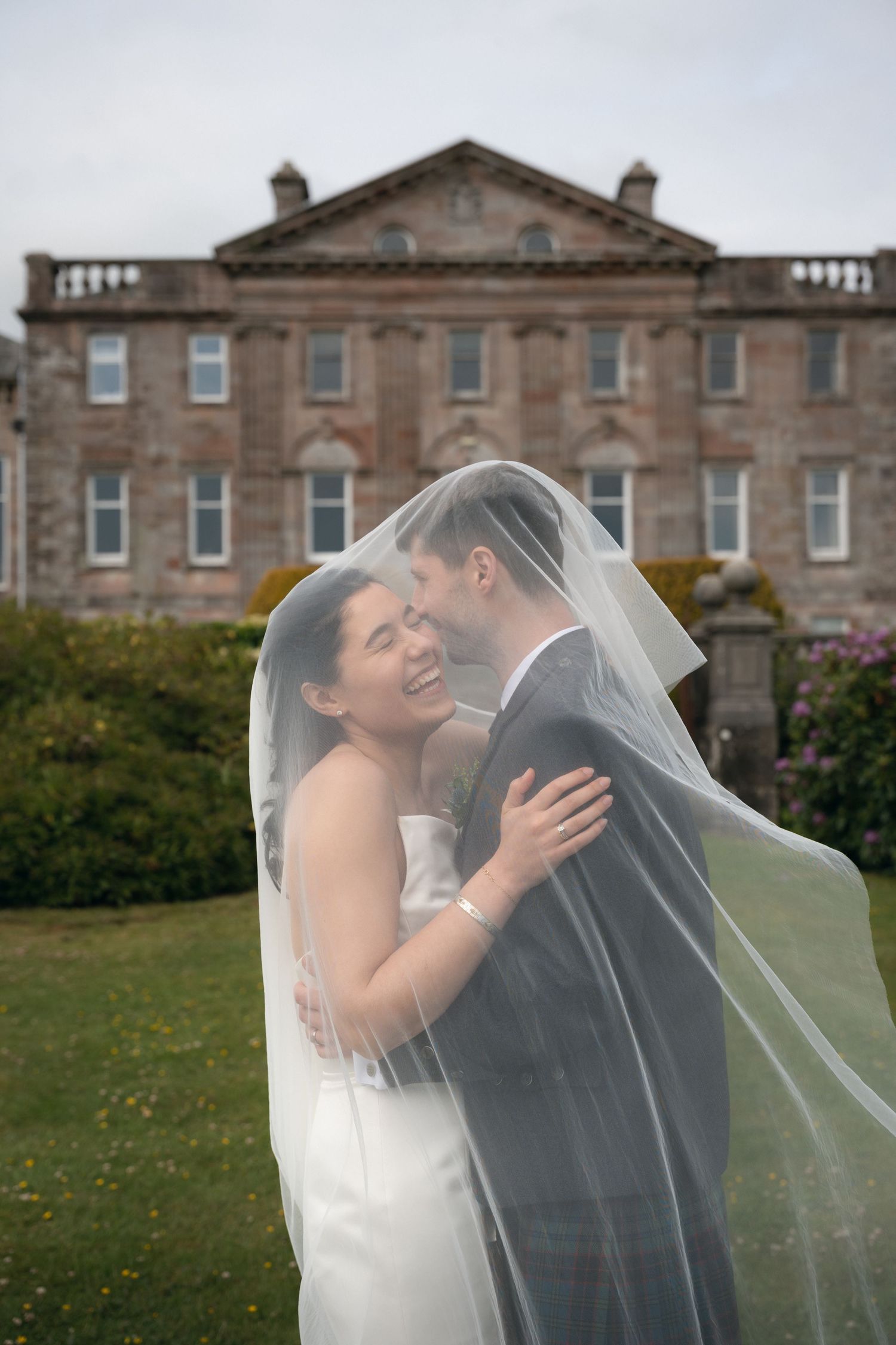 A romantic wedding couple embraces outdoors with their veil flowing in front of a historic stone mansion.