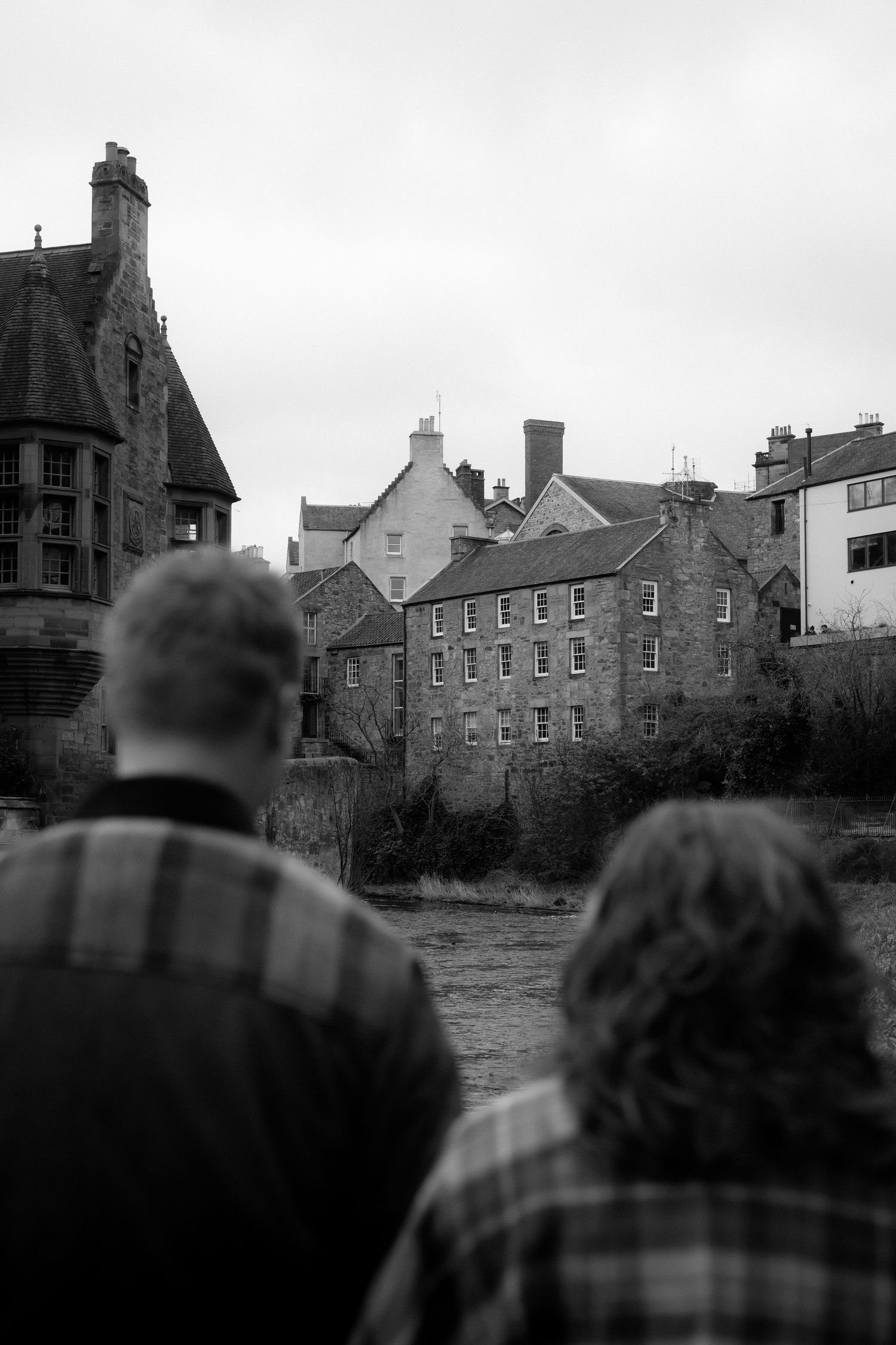 Black and white photo of two people viewing historic stone buildings along a river in a quaint European town.