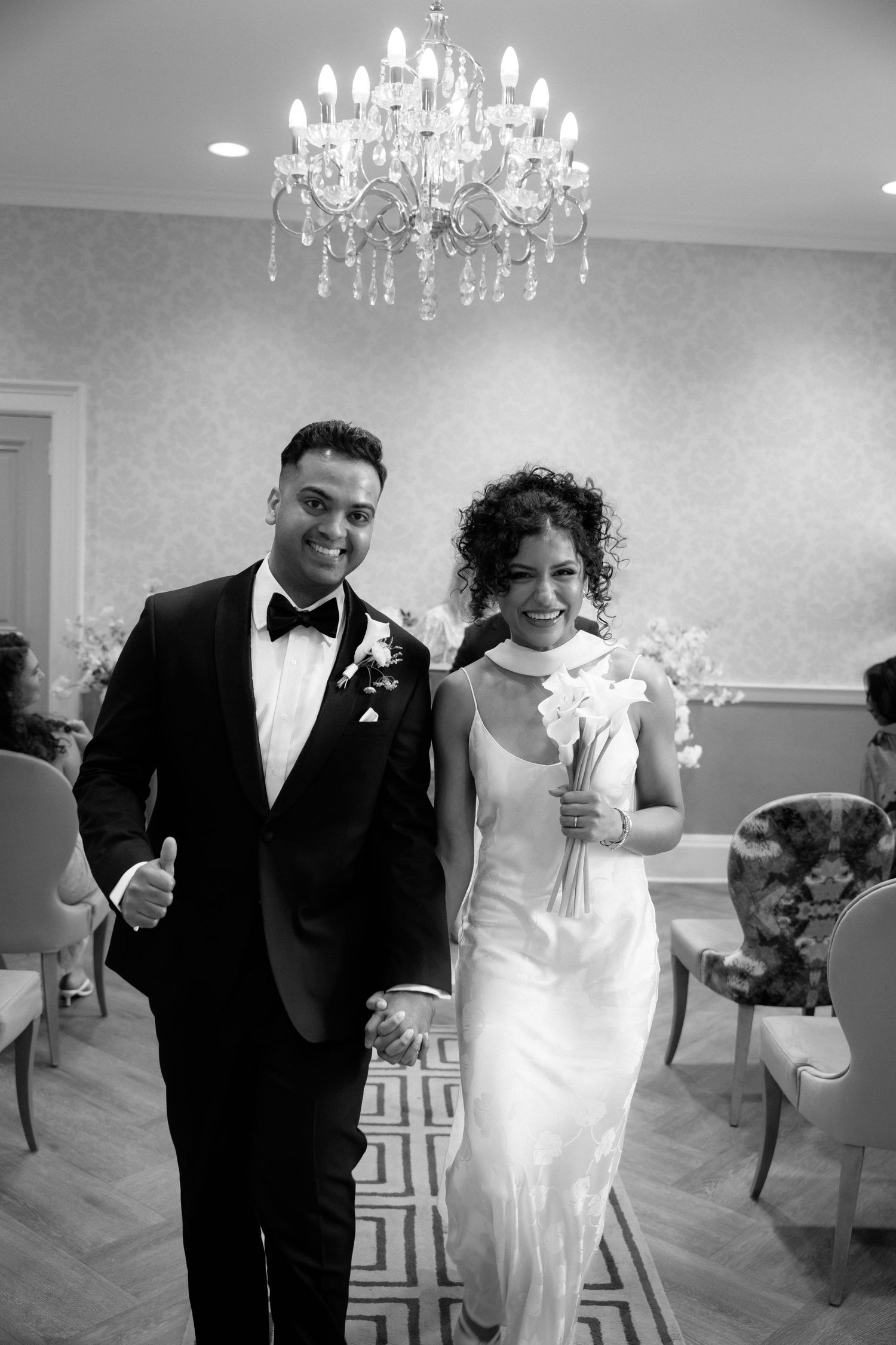 A couple in formal wedding attire dances and celebrates during their reception under elegant crystal chandeliers.