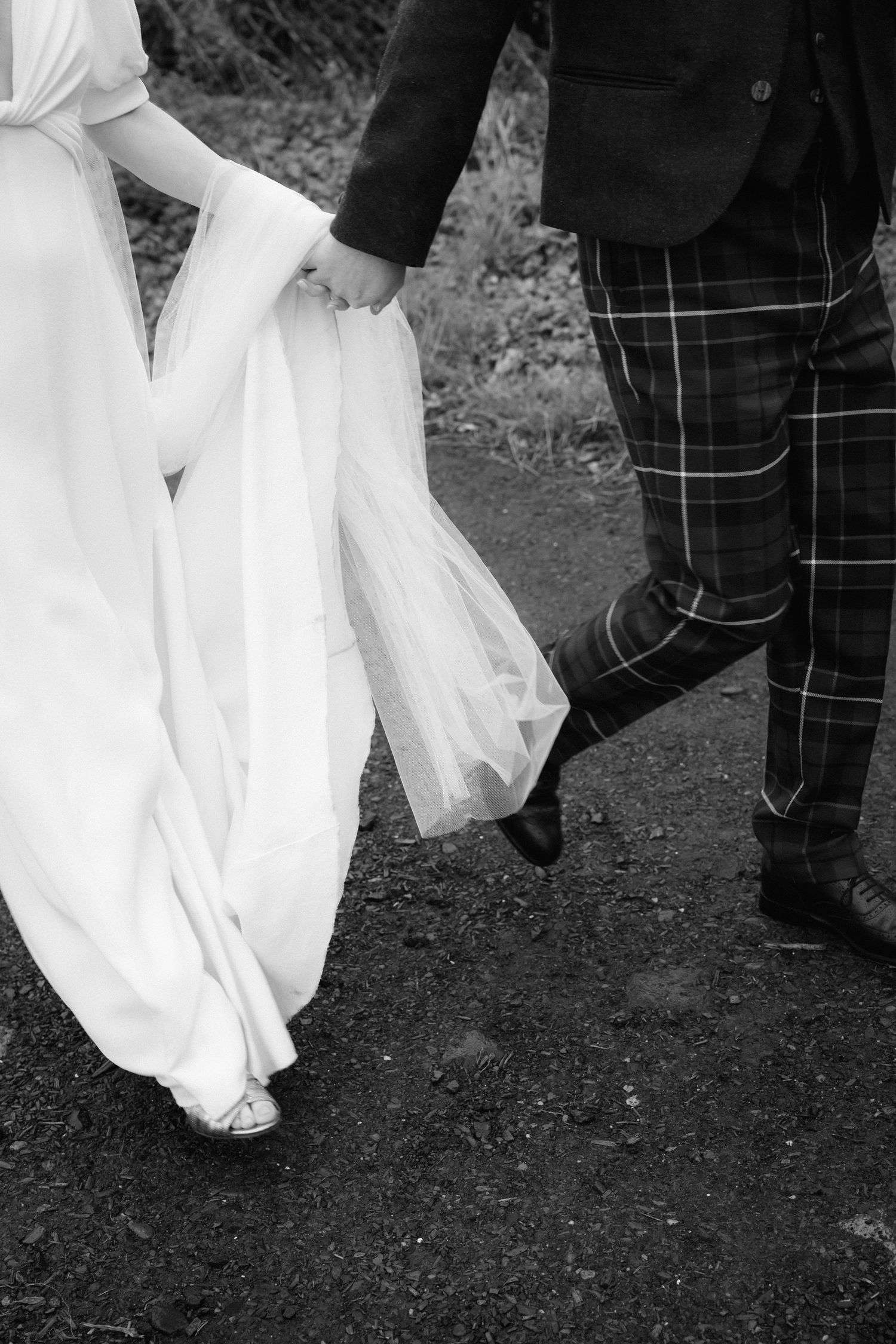 Black and white photo showing the hem of a white wedding dress next to legs wearing plaid pants.