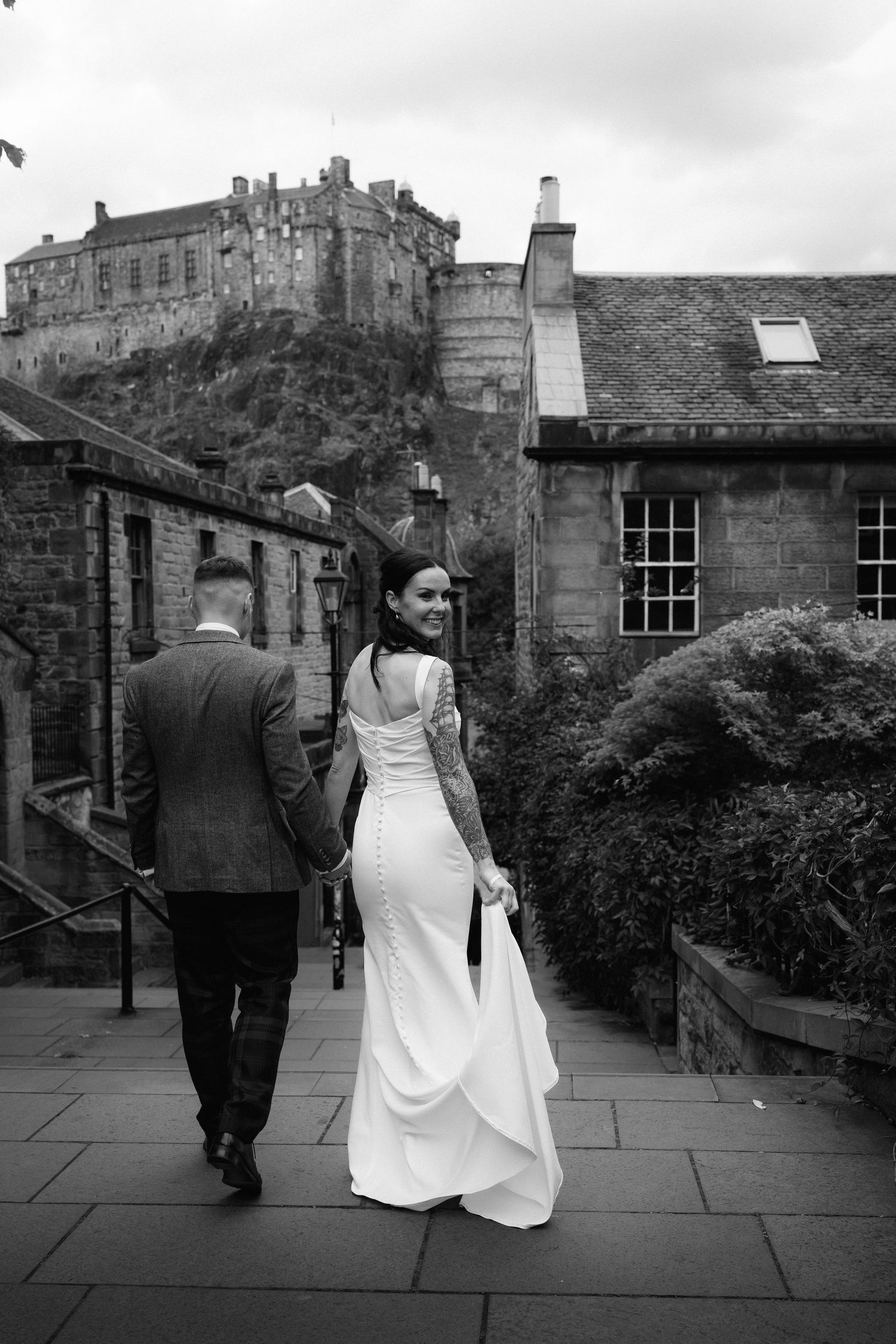 A wedding couple walks along a stone path with Edinburgh Castle looming dramatically in the background.