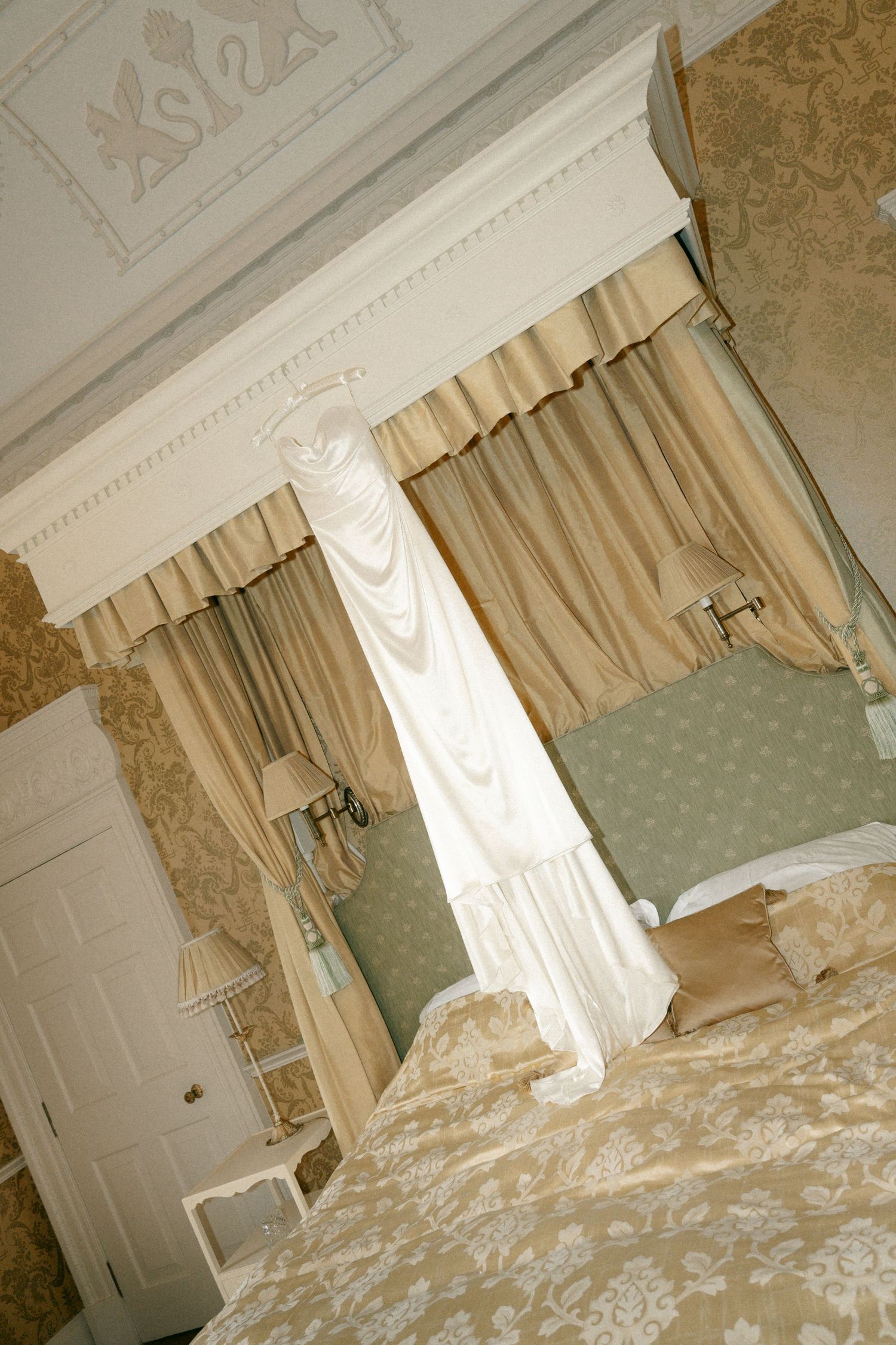 Elegant bedroom interior with ornate wallpaper, white crown molding, and cream colored curtains filtering natural light.