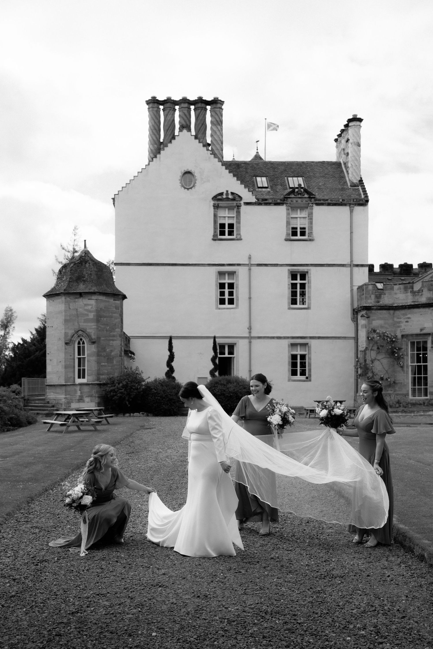 Wedding party arranges bridal veil on lawn outside elegant historic castle with turret and chimneys in black and white photo.