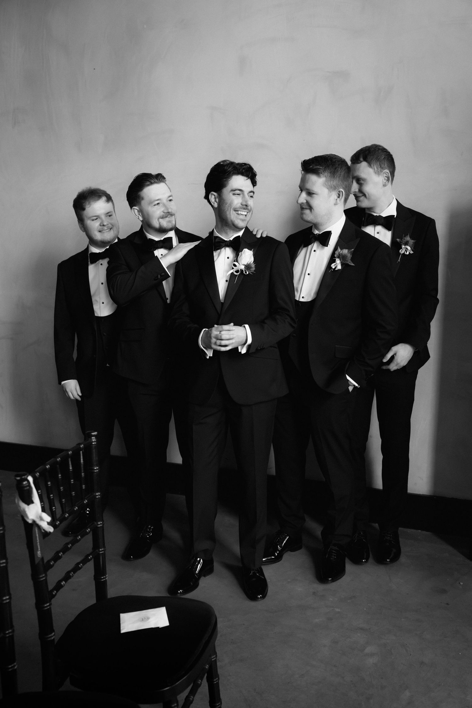 Black and white photo of a groom and four groomsmen in tuxedos laughing together before a wedding ceremony.