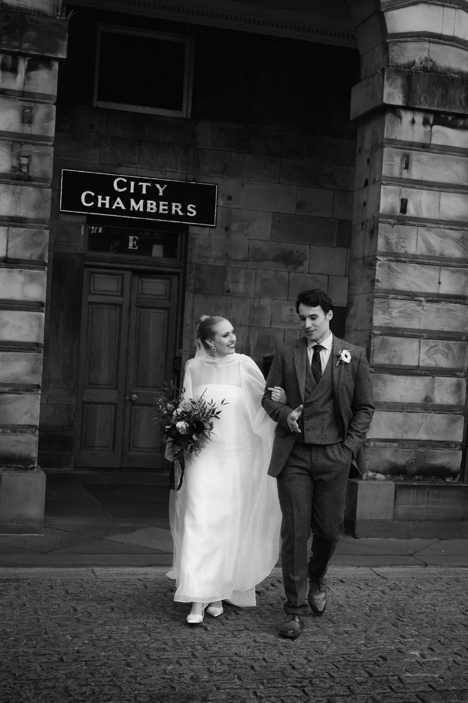 A wedding couple walks together on cobblestones outside City Chambers in a black and white artistic sequence.