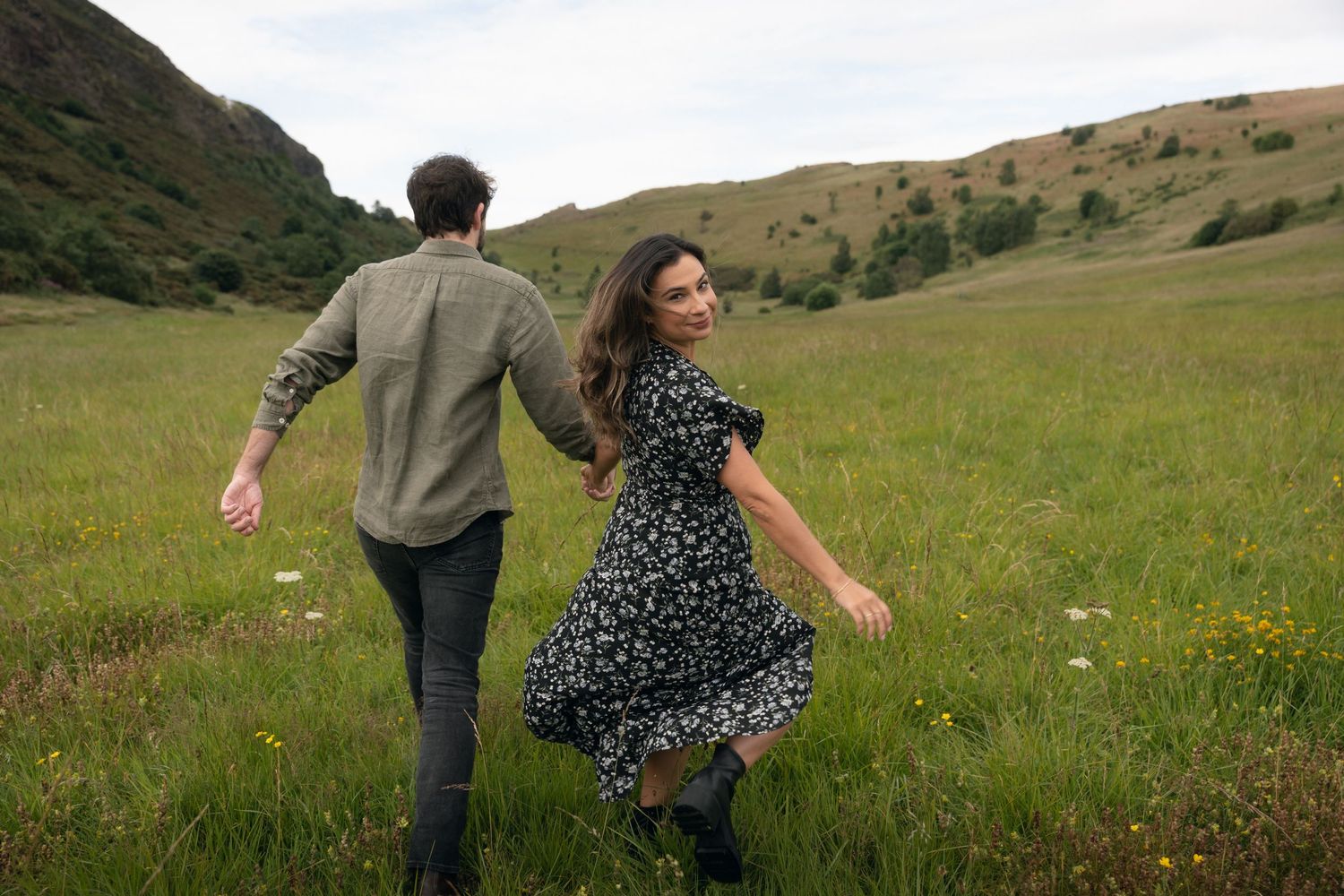 Two people run through a green meadow holding hands with mountains in the background on a cloudy day.