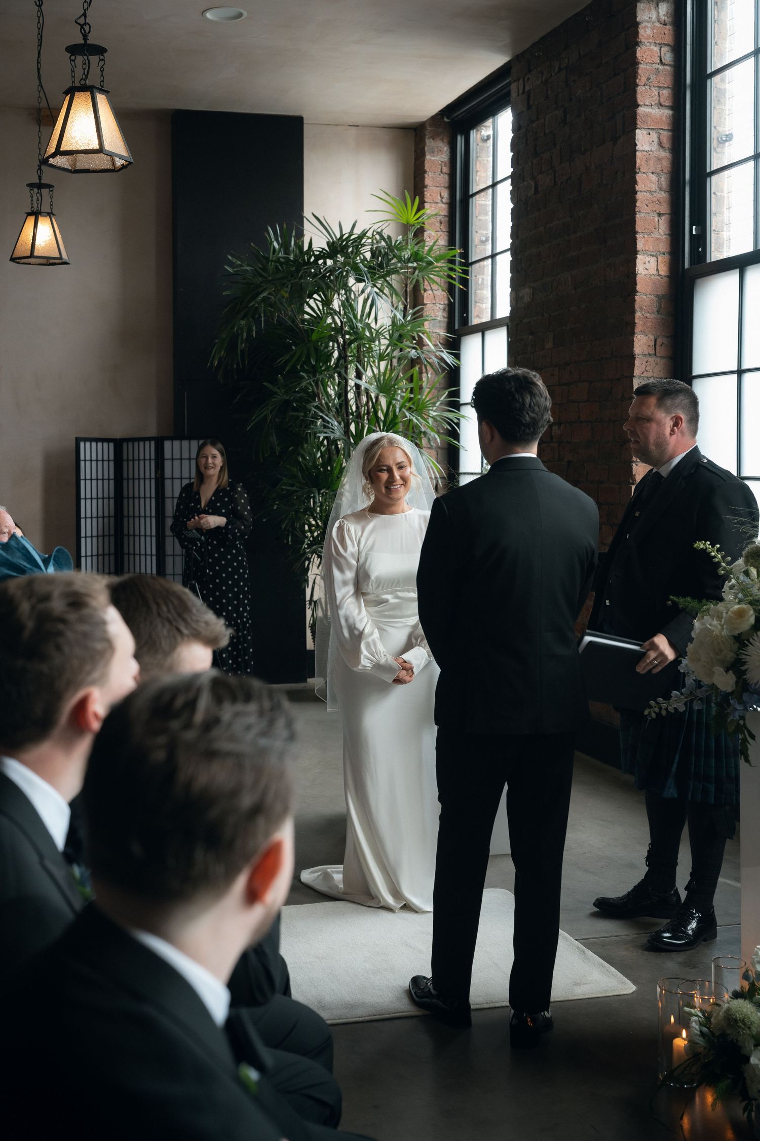 Bride in elegant white gown smiling at groom during indoor wedding ceremony with brick walls and tropical plants.