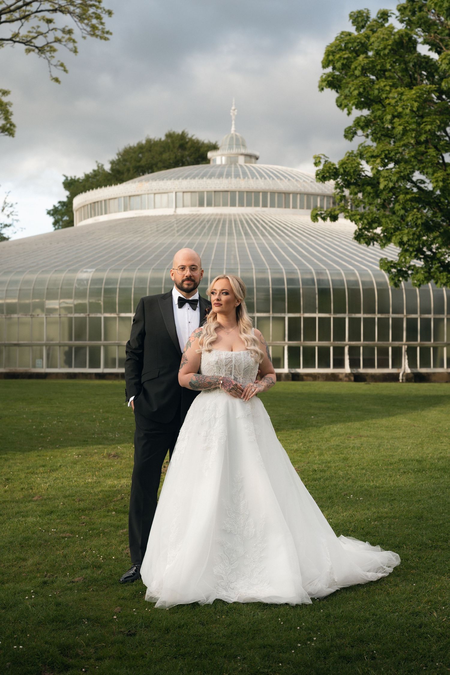 Bride in white ballgown and groom in black tuxedo posing in front of a glass greenhouse on their wedding day.