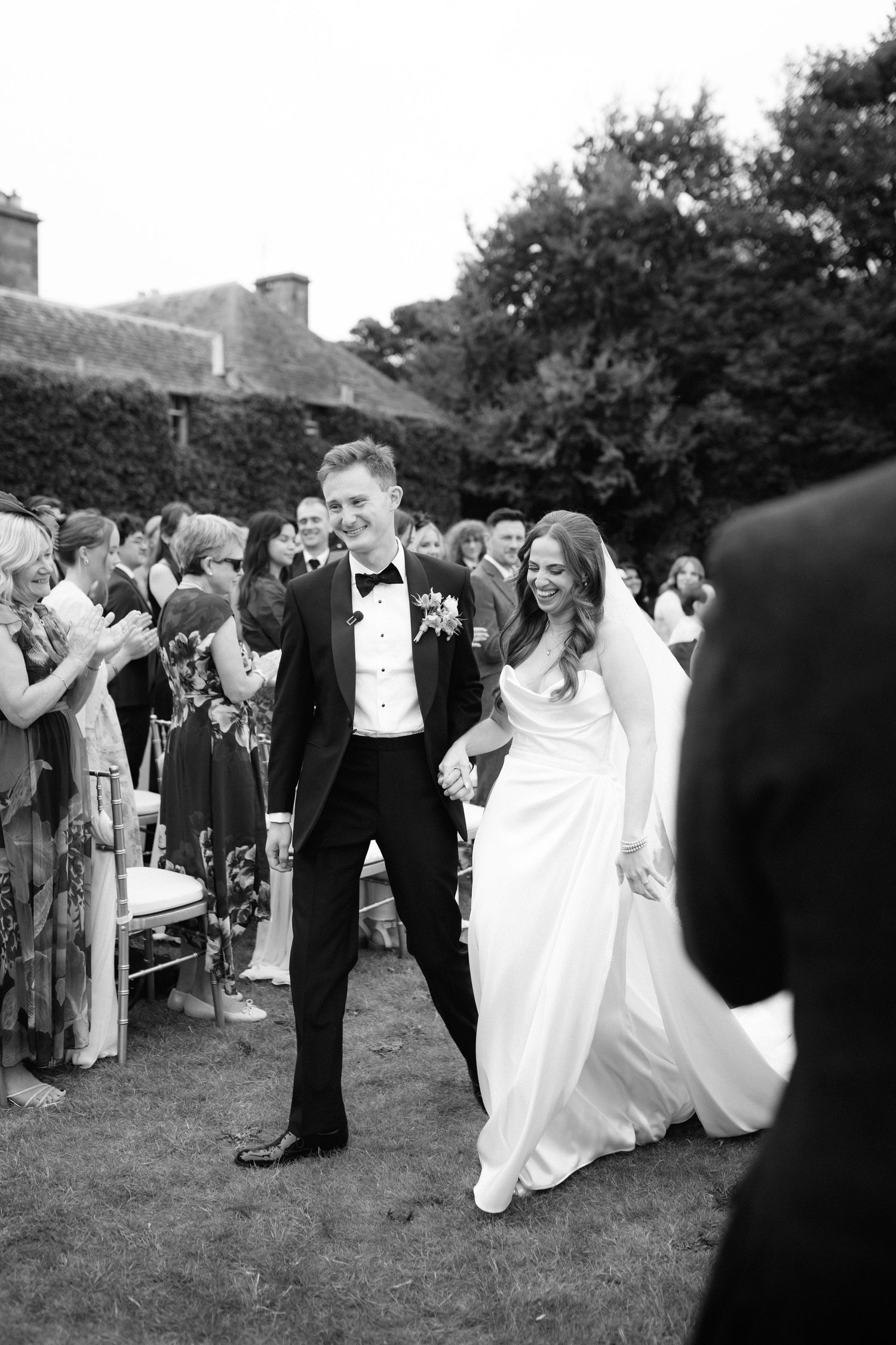 A black and white wedding photo of a joyful couple walking through guests on a garden lawn during their celebration.