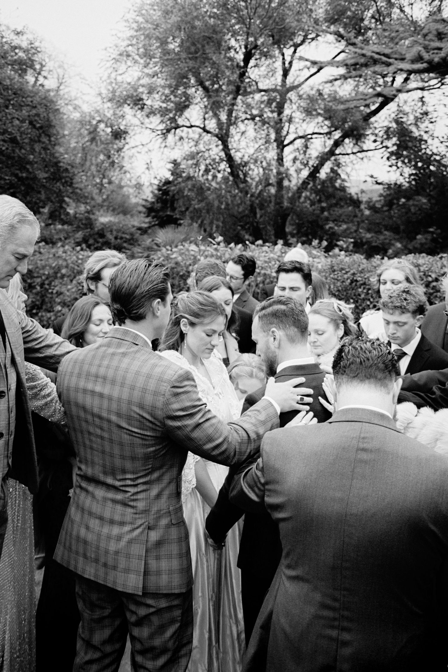 Black and white photo of wedding guests gathered outdoors under trees during a celebration.