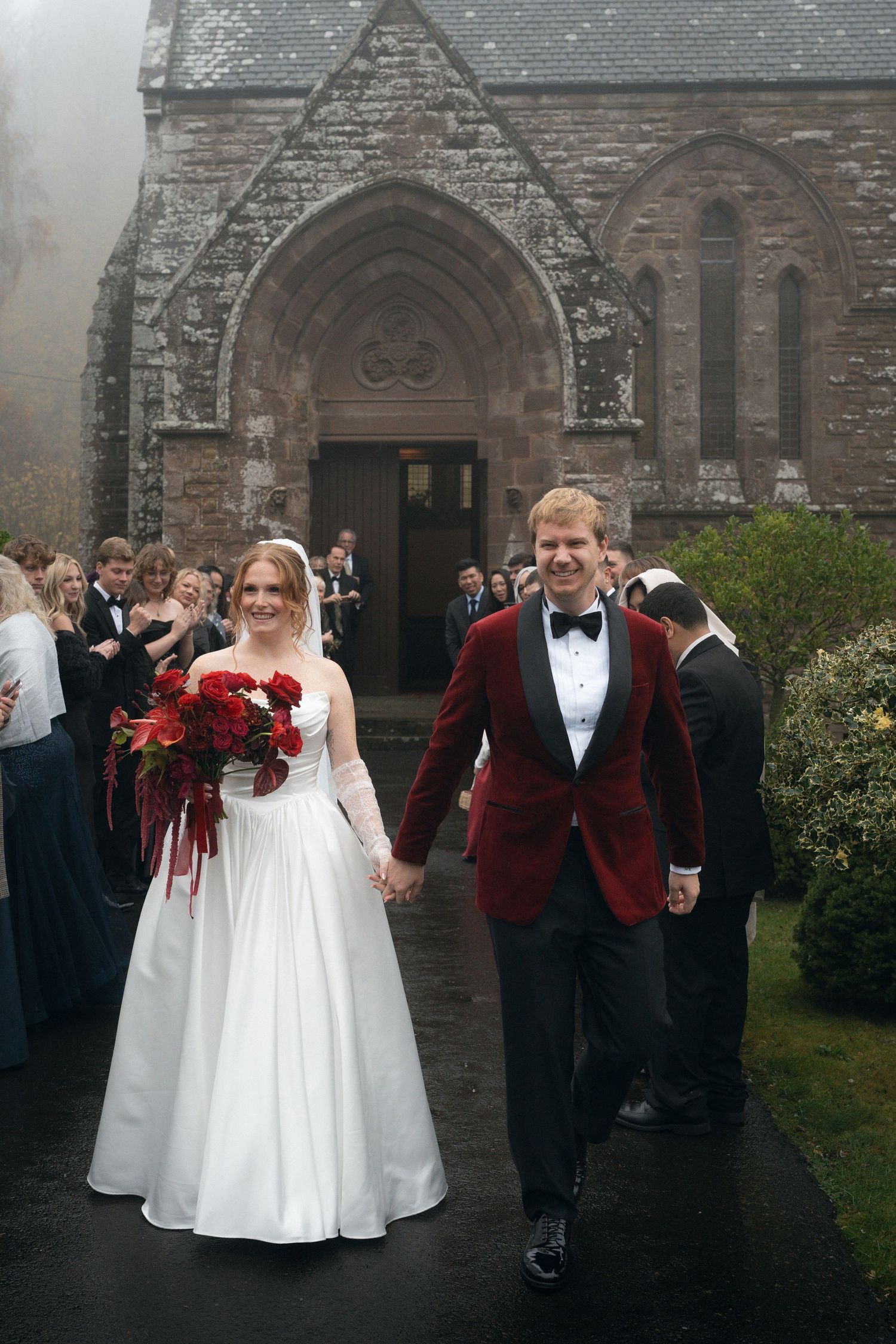 Wedding party poses outside a stone church with bridesmaids in burgundy dresses and groomsmen in matching suits.