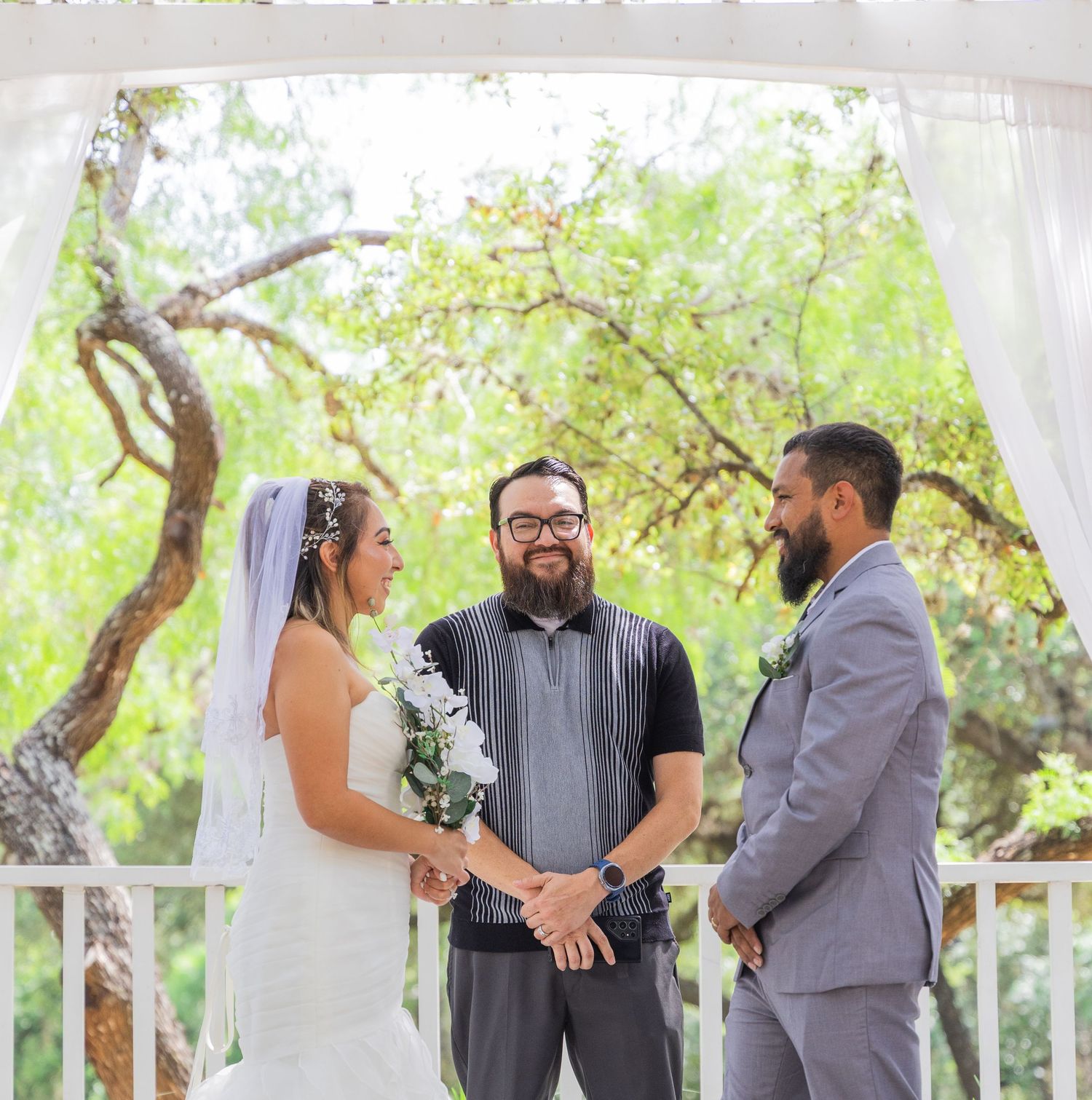 Bride and groom exchange vows with bearded officiant under white draped pergola at outdoor wedding.
