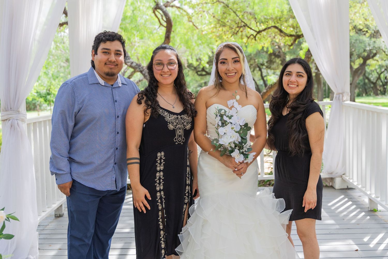 Bride in white strapless gown holds white bouquet posing with three guests under wedding arch.
