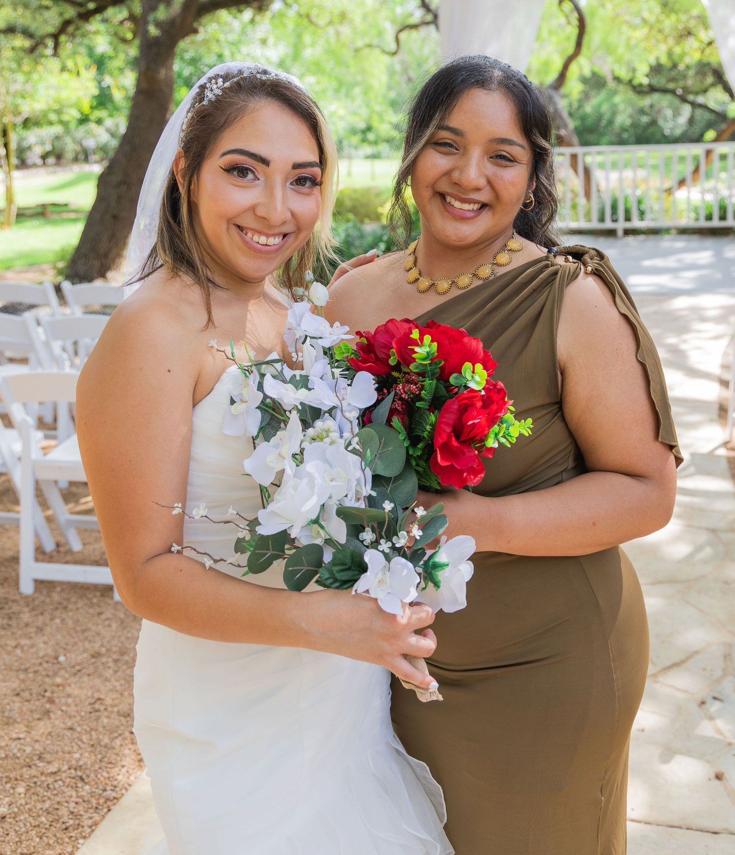 Bride in white strapless gown and bridesmaid in olive dress holding bouquets outdoors.
