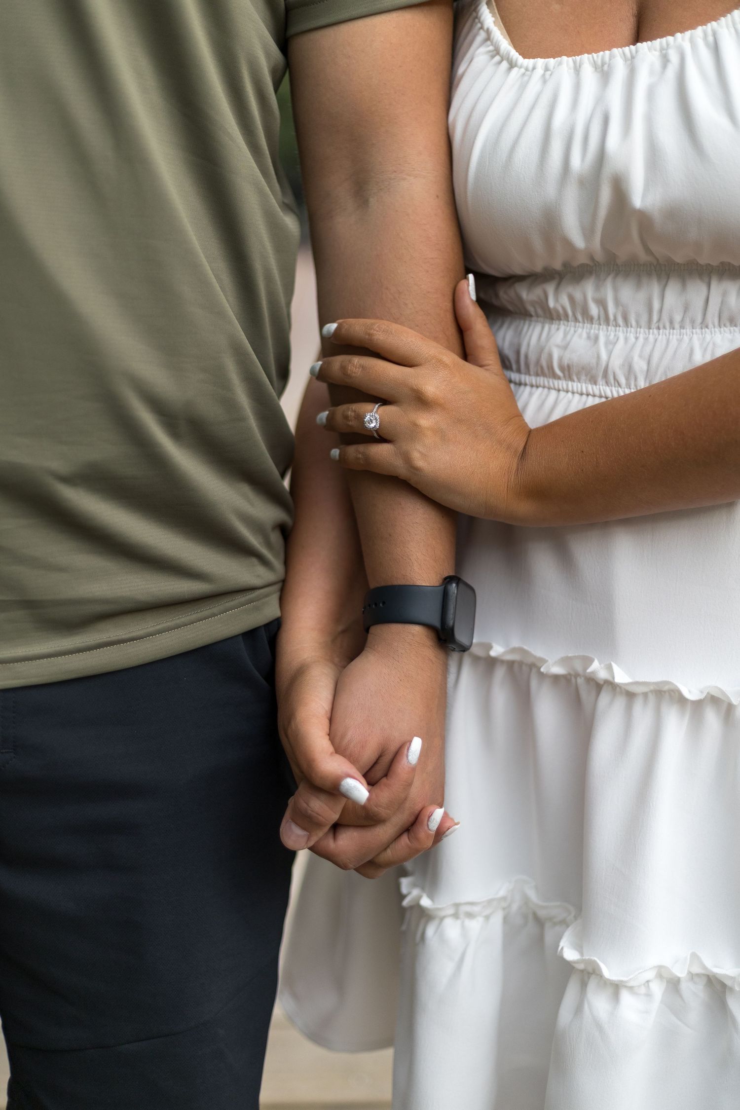 Close-up of interracial couple holding hands, woman in white dress showing engagement ring and fitness tracker.