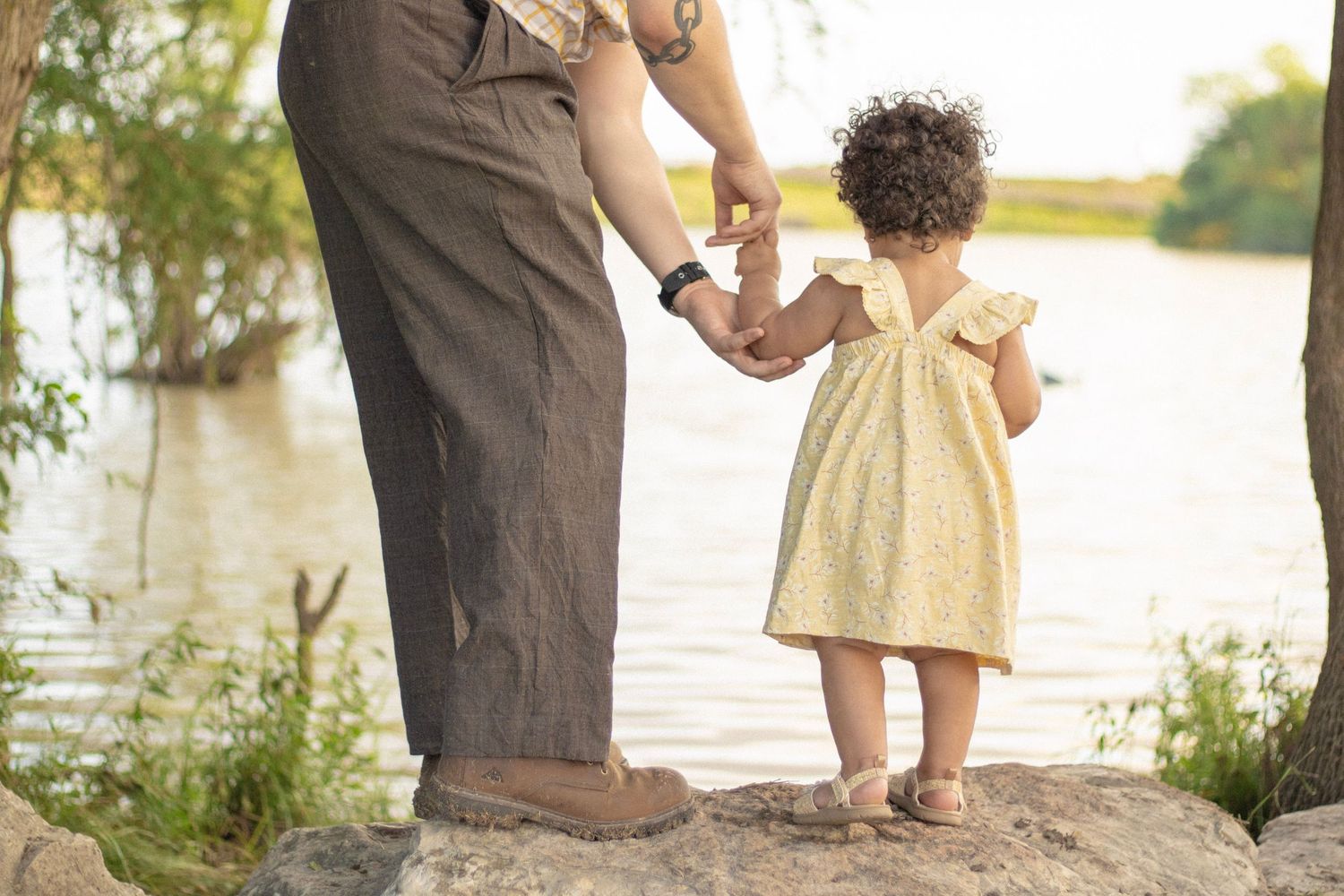 Toddler girl in yellow dress holds adult hand while standing on rocks beside a peaceful lake.