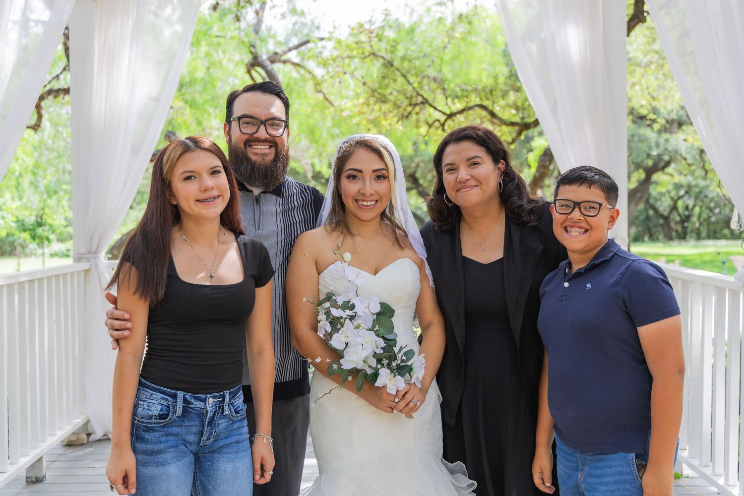 Bride in white gown holds bouquet surrounded by family at outdoor wedding ceremony under white arbor.