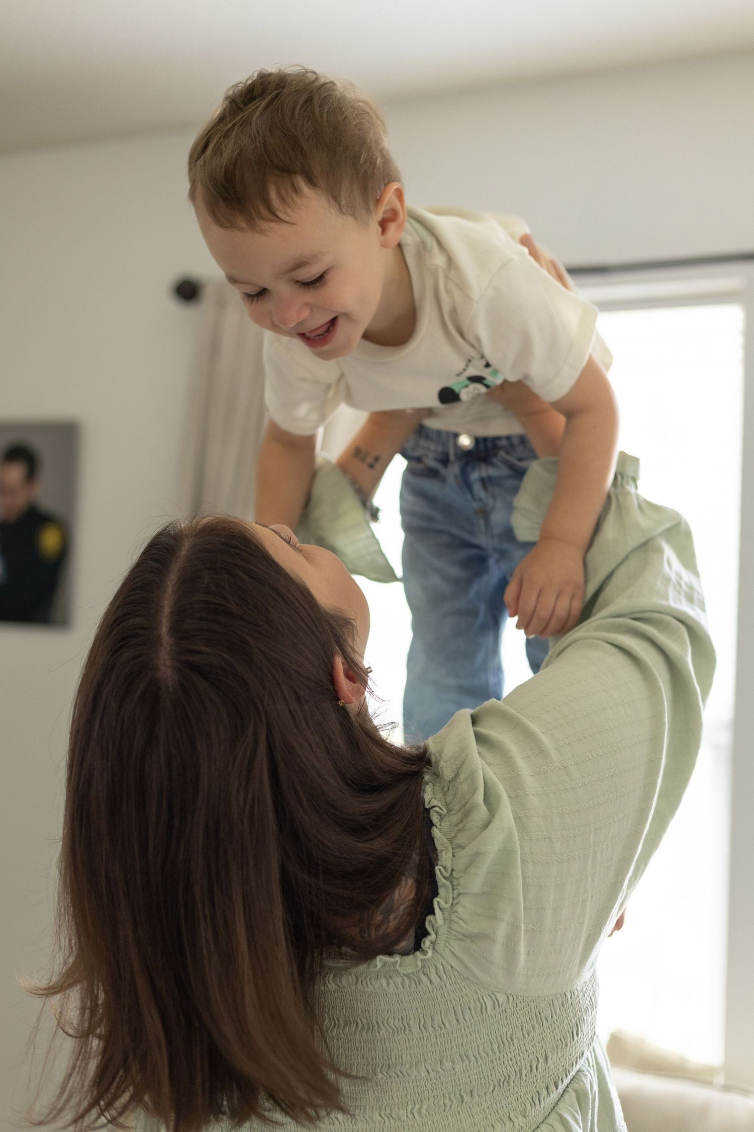 Joyful mother in green top lifting laughing toddler boy high in bright living room.