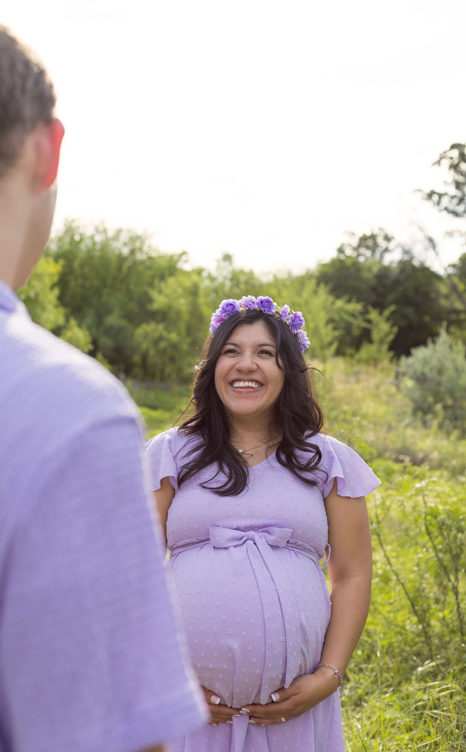 Smiling pregnant woman in purple floral crown and lavender dress during outdoor maternity shoot.