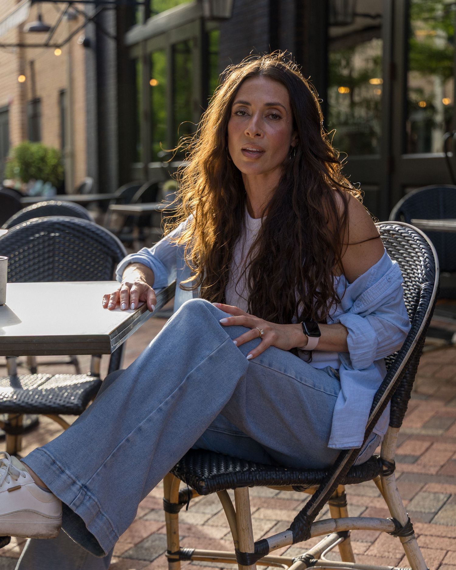 Woman with long wavy hair sits relaxed at outdoor cafe patio table.