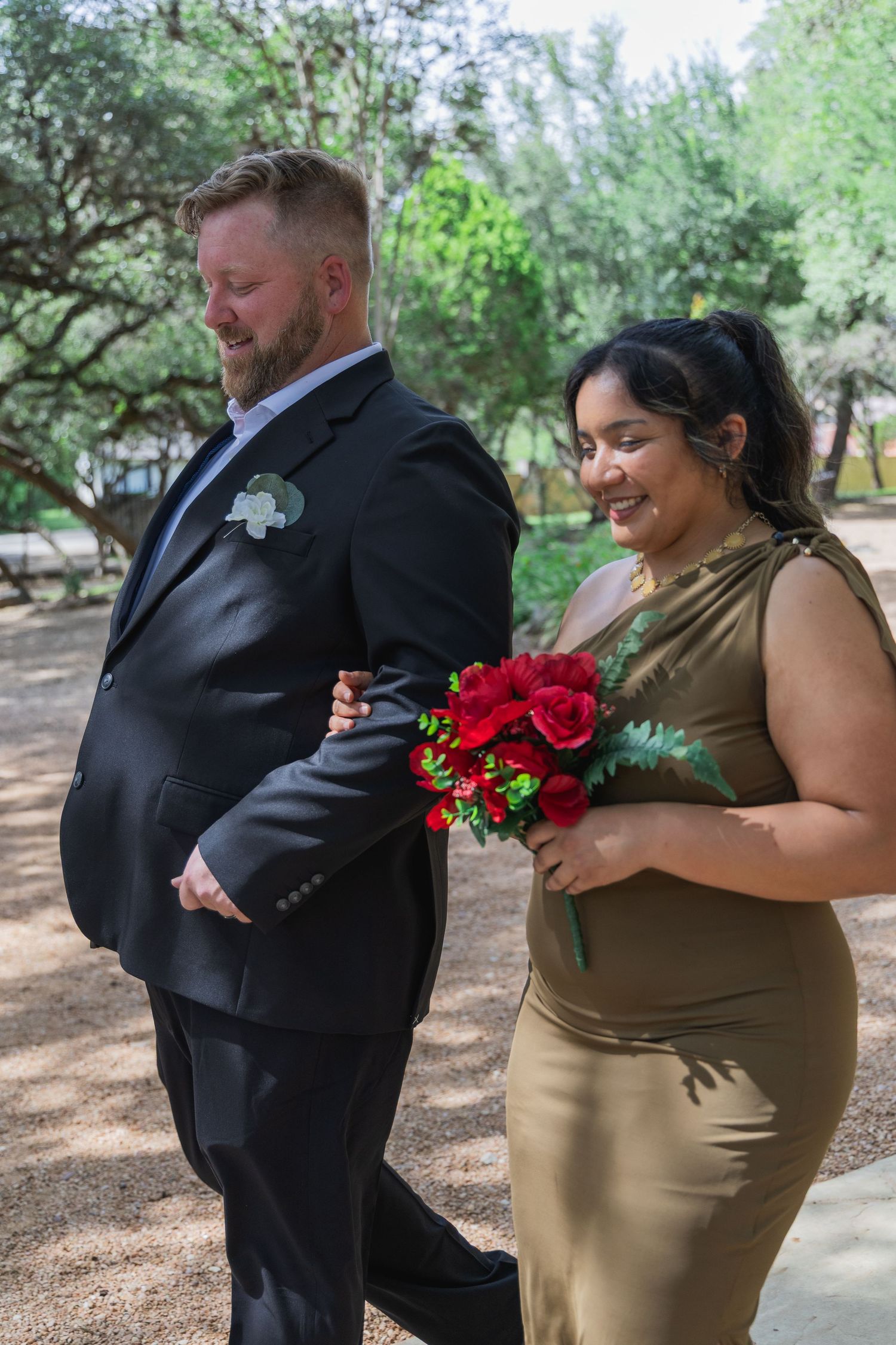 Groomsman in black suit walks with bridesmaid in gold dress holding red rose bouquet at outdoor wedding.
