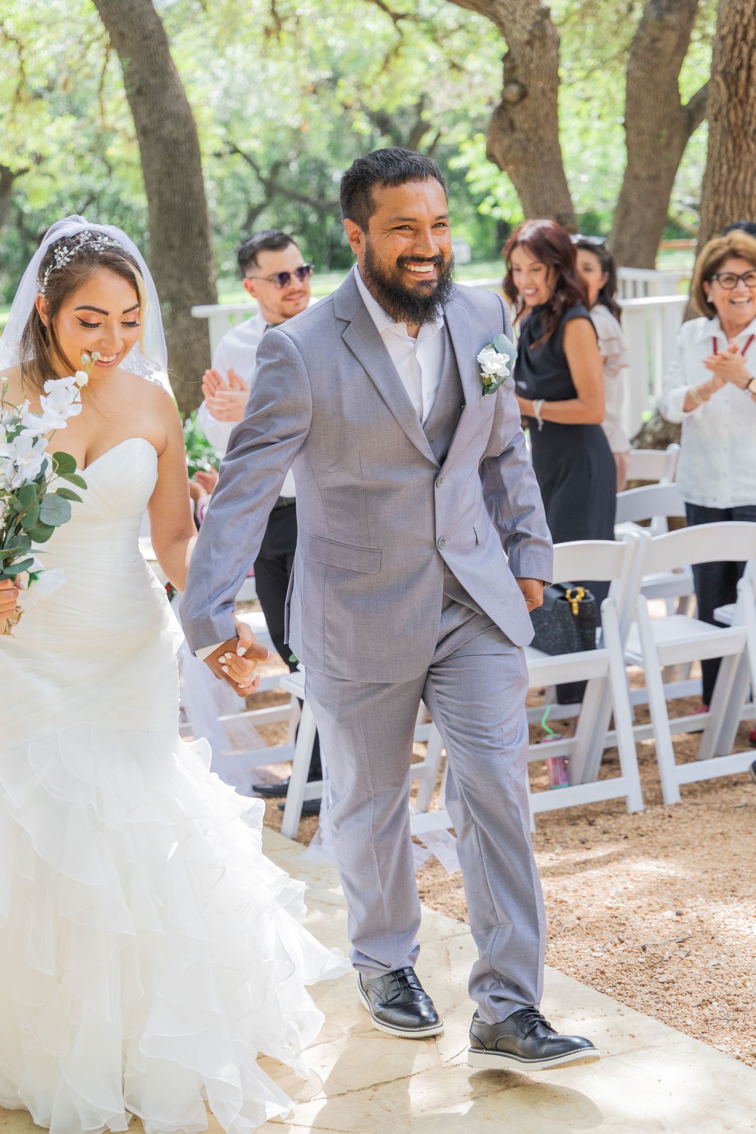 Joyful groom in gray suit walks bride down outdoor wedding aisle surrounded by guests under trees.