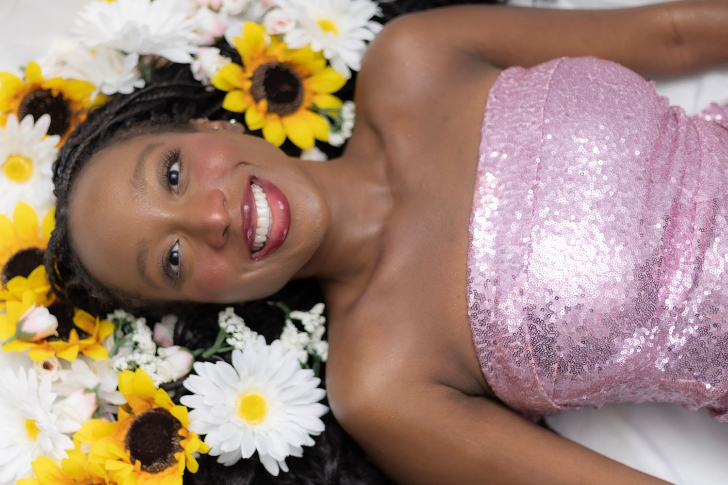 Joyful young Black woman in pink sequin top lying among sunflowers and white daisies smiling.