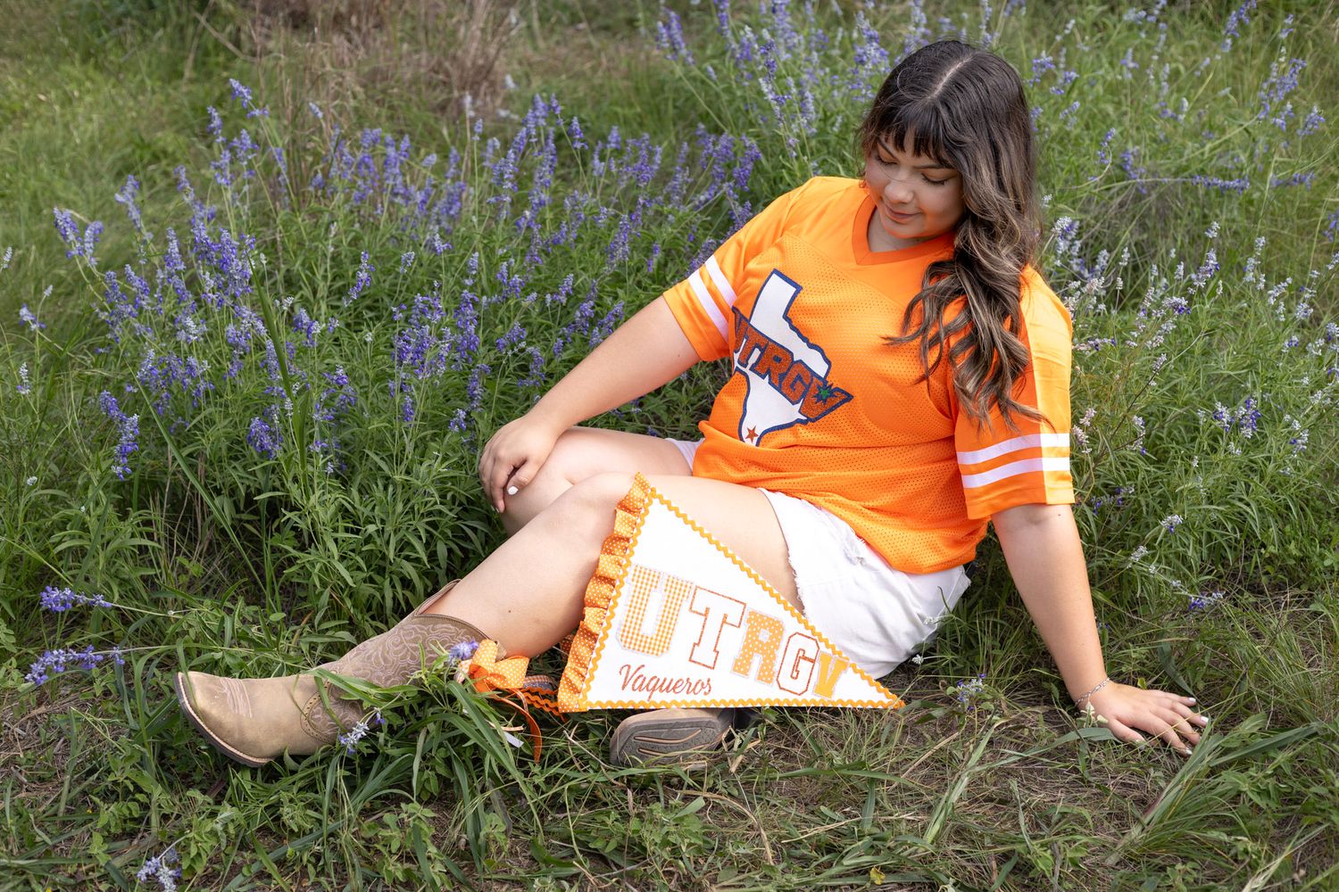 Young woman in orange UTRGV jersey sits in bluebonnet field holding pennant flag.