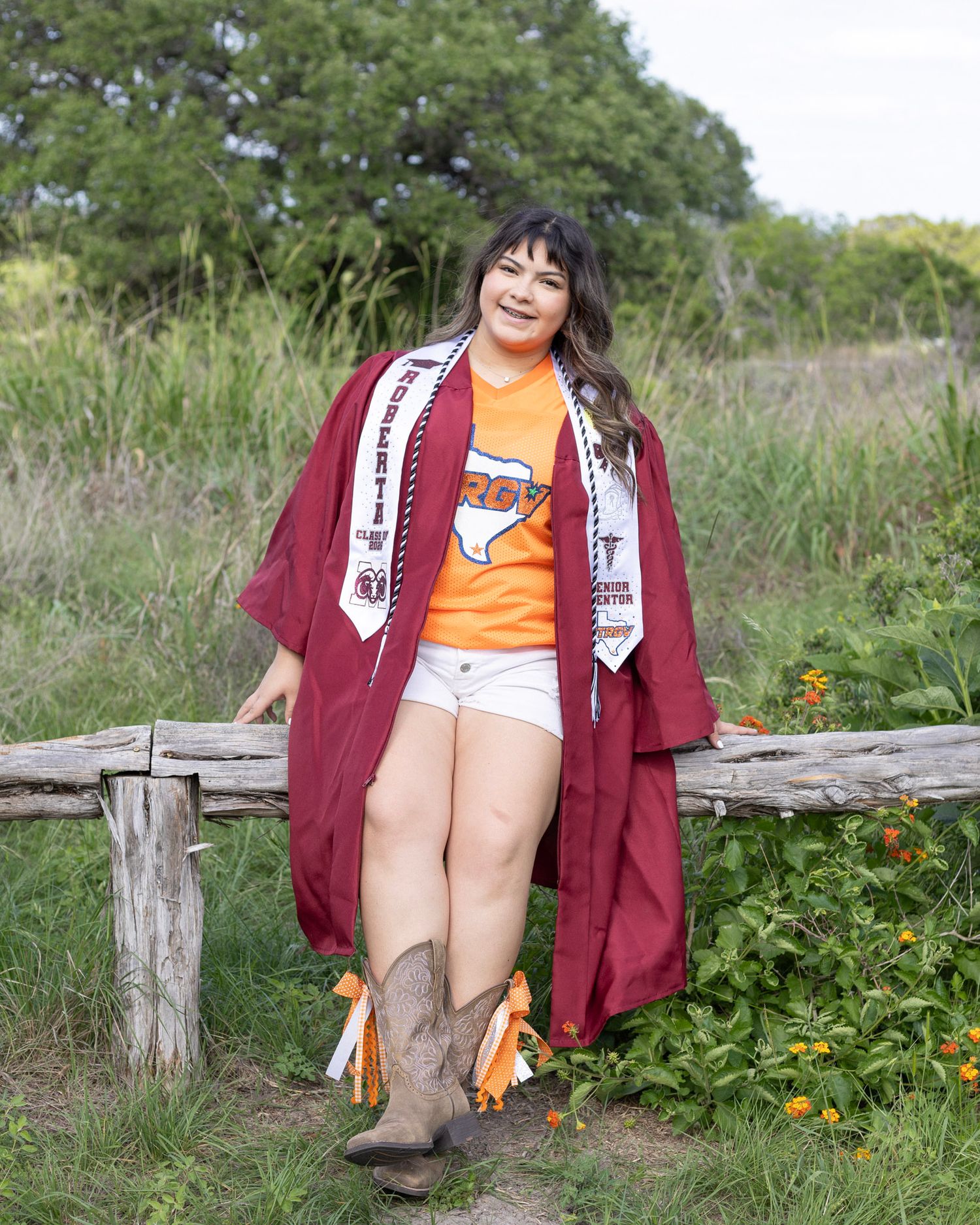 Graduate in maroon cap and gown with orange jersey poses on rustic fence outdoors.