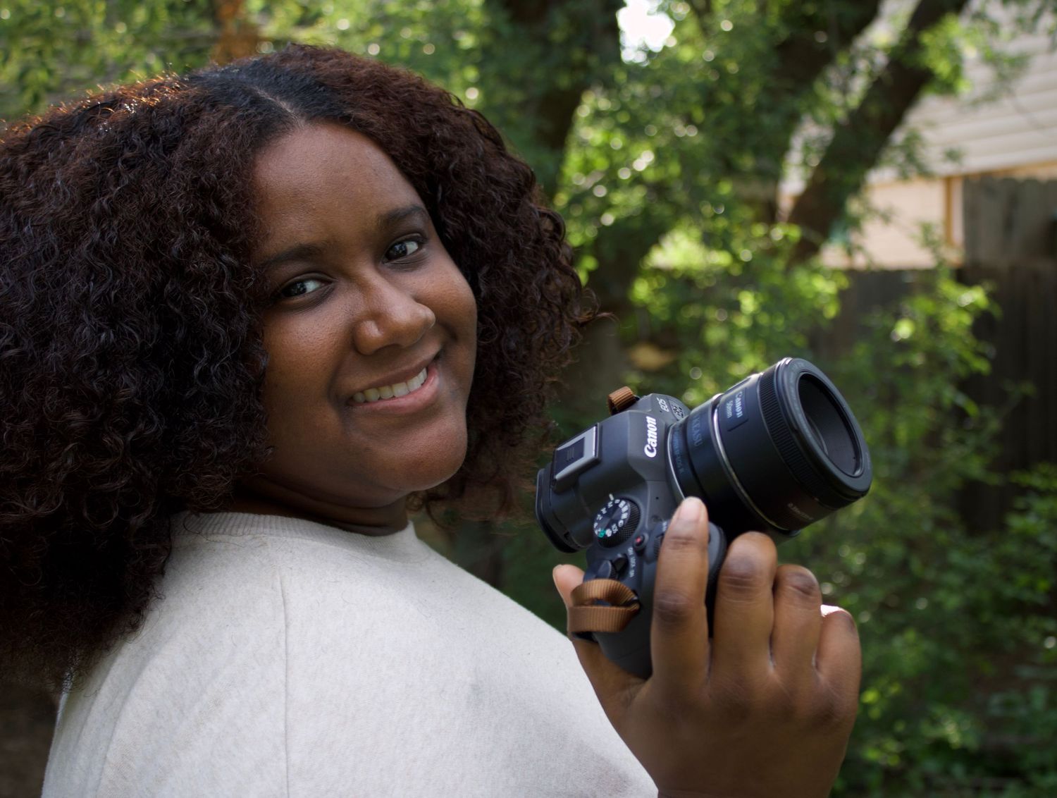 Smiling woman with curly hair holding a DSLR camera outdoors in green garden setting.